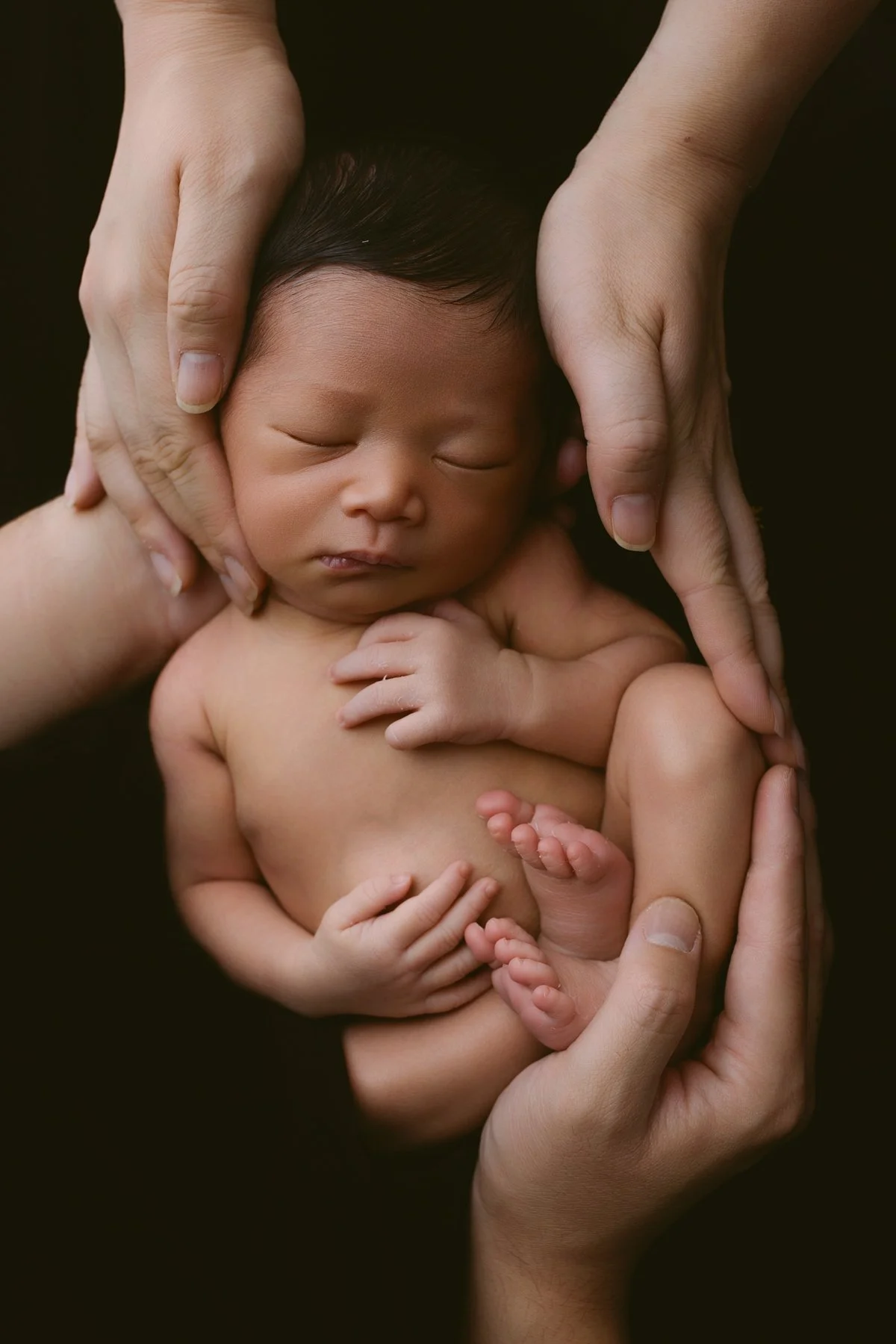 A newborn baby peacefully sleeping, surrounded by parents hands gently cradling the baby.