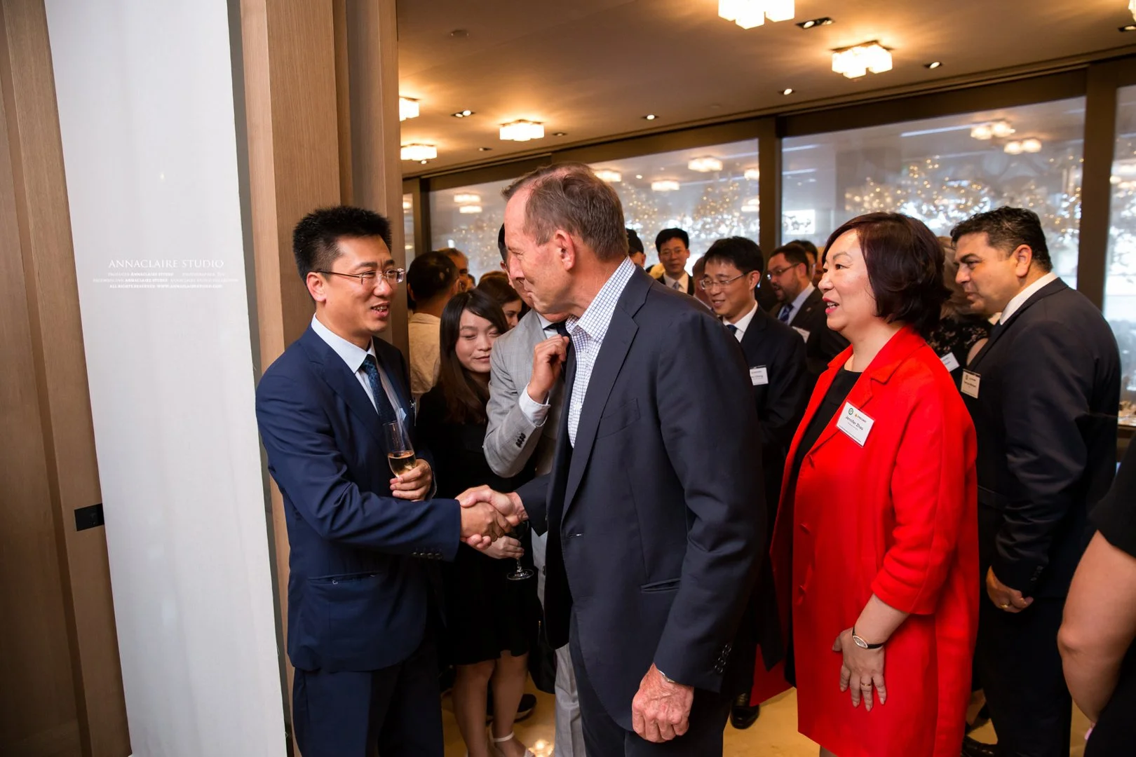 A group of business professionals at a networking event. A man in a gray suit is shaking hands with a man in a blue suit and glasses, holding a glass of champagne. Several other people are engaged in conversations, with one woman in a bright red blaz