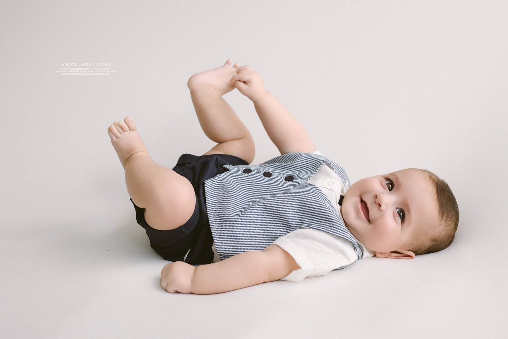 A baby lying on the floor on a light background, smiling and looking at the camera, dressed in a white shirt with a blue vest and black shorts.