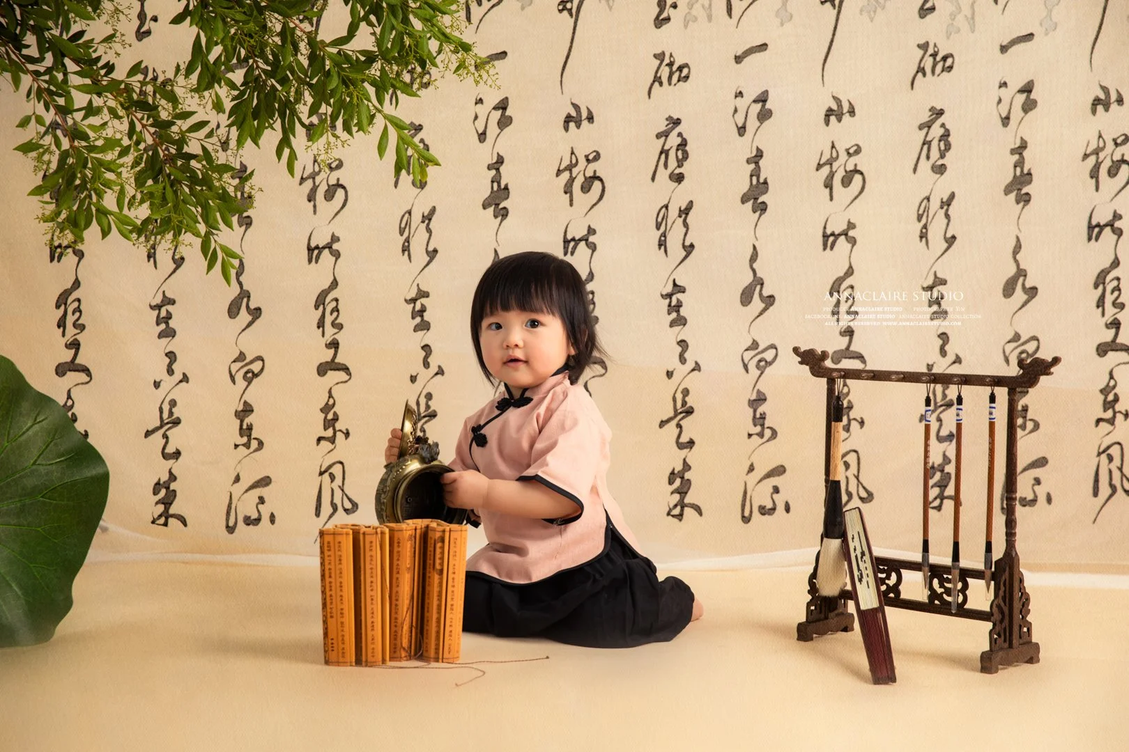 the 1 year old baby girl dressed in traditional Asian attire sitting on the floor surrounded by books and musical instruments, with calligraphy in the background and decorative plants nearby.