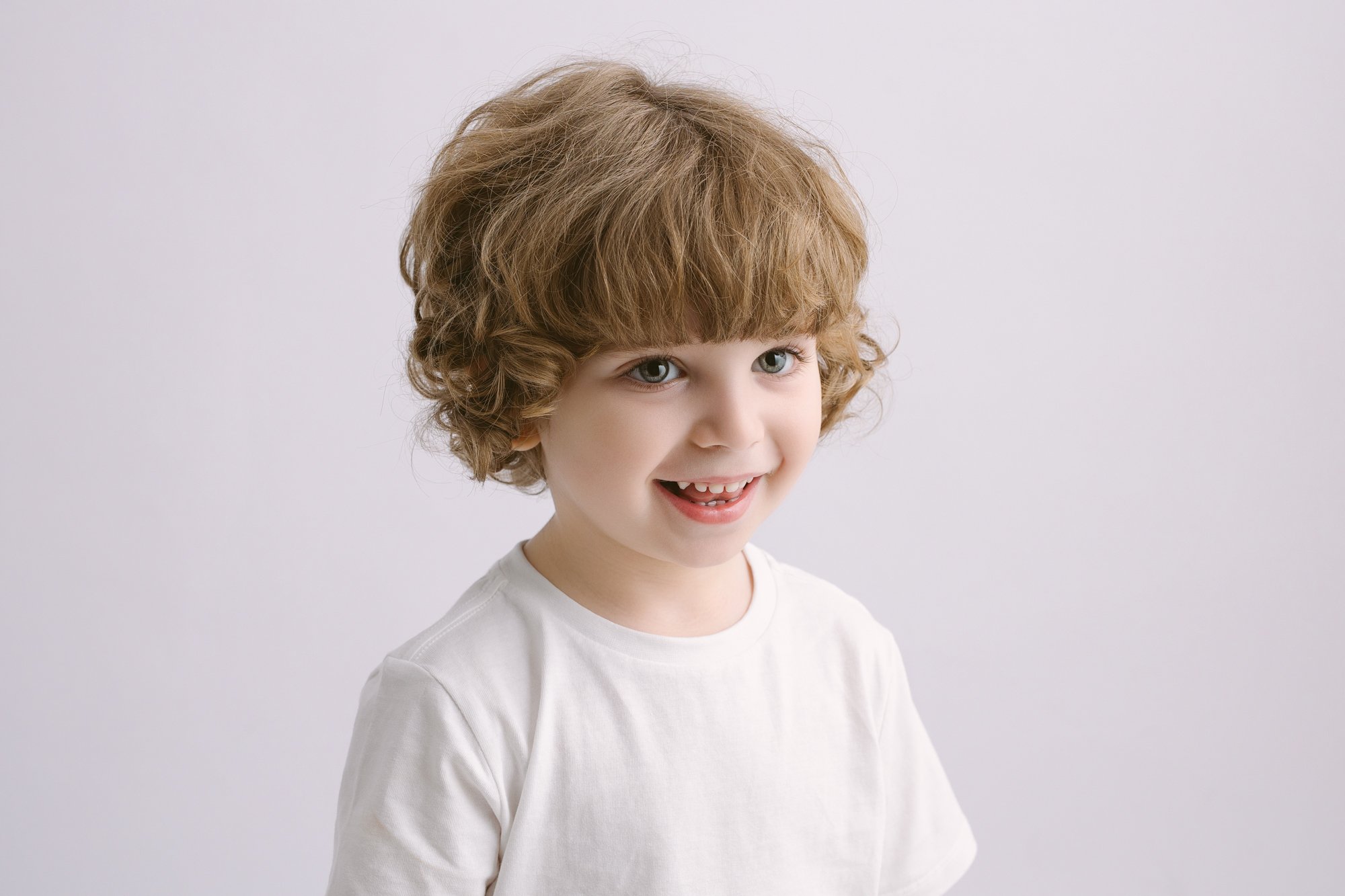 A young boy with curly light brown hair, blue eyes, and a wide smile showing some missing teeth, wearing a plain white t-shirt against a neutral background.