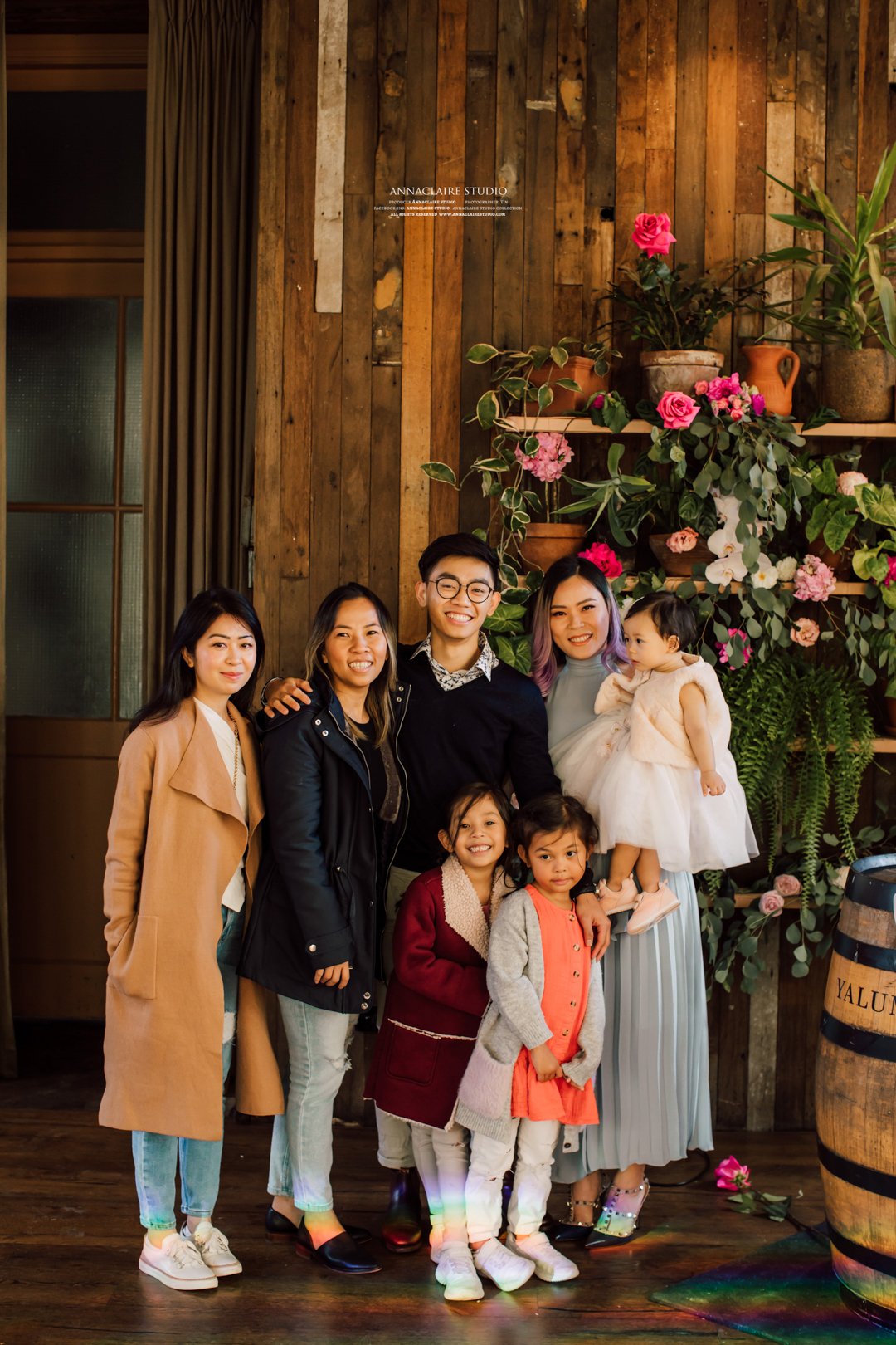 A group of eight people, including five children and three adults, standing together indoors in front of a wooden wall with pink and white flowers, smiling for a photo.