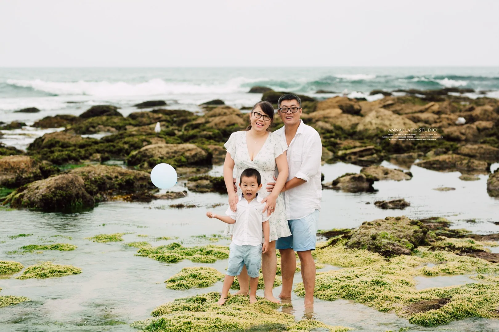 A family of three standing on a rocky, mossy beach with the ocean in the background during overcast weather. The mother, father, and young son are smiling, and the son is holding a balloon.