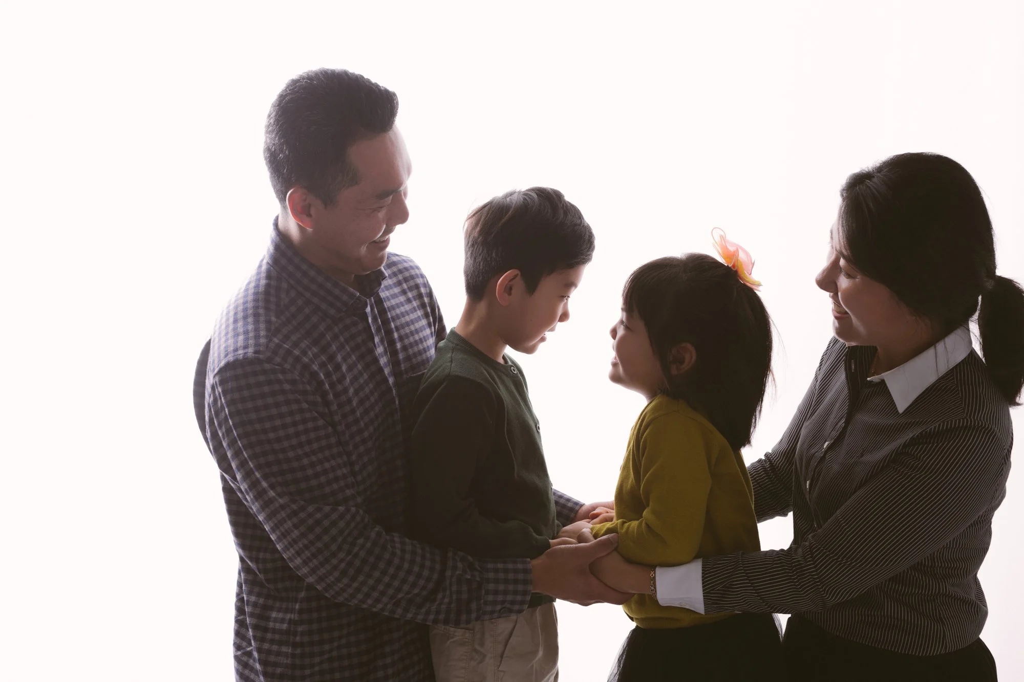 Family of four smiling and holding hands, with the children facing each other and the parents holding the children's hands in the middle, against a plain white background.
