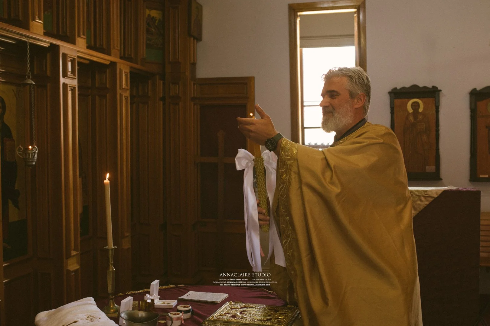 A man in gold religious robes performing a ritual inside a church, holding a lit candle with a white ribbon, surrounded by religious icons and candles.