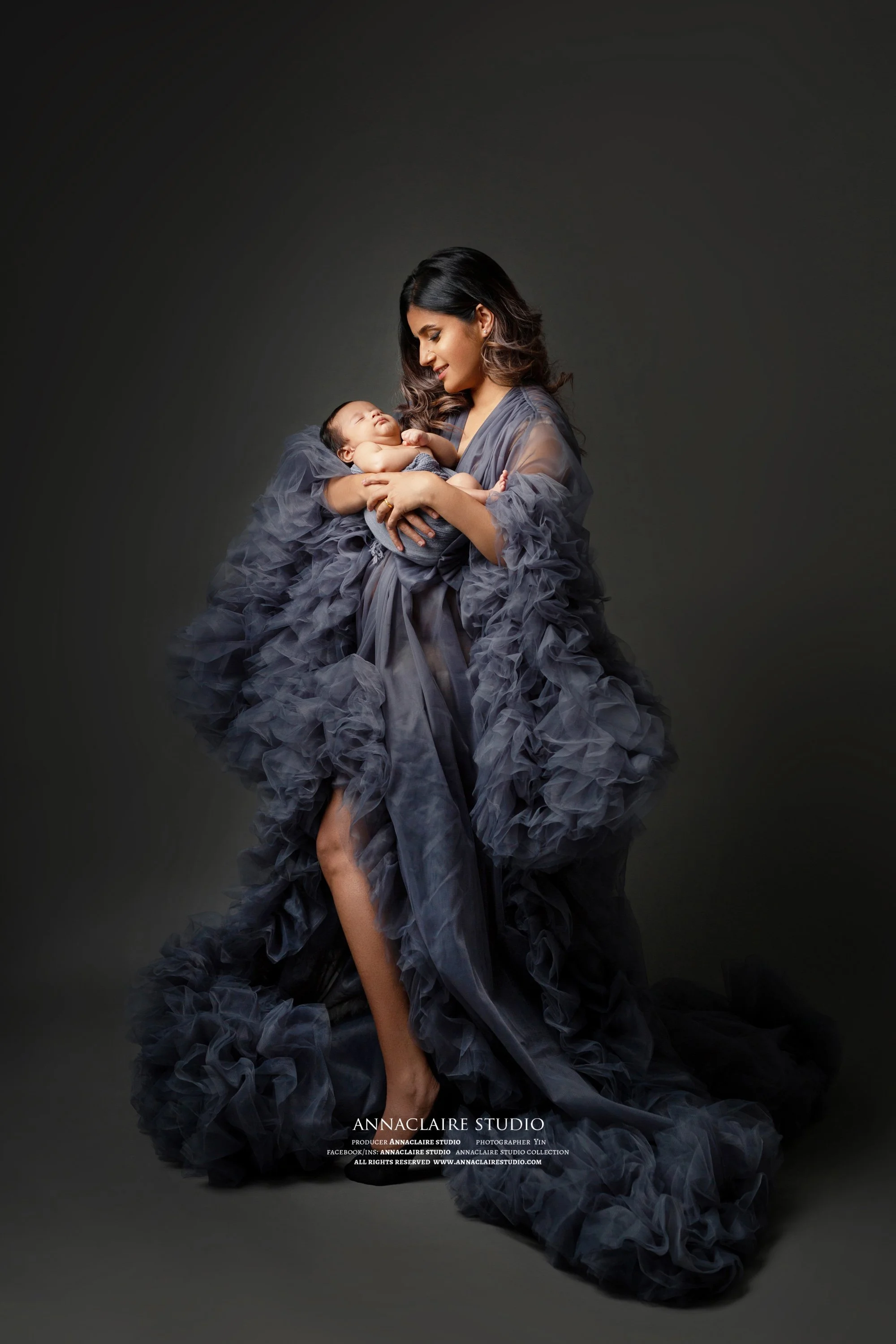 A woman in an elegant gray, ruffled gown holding a sleeping baby against a dark background.