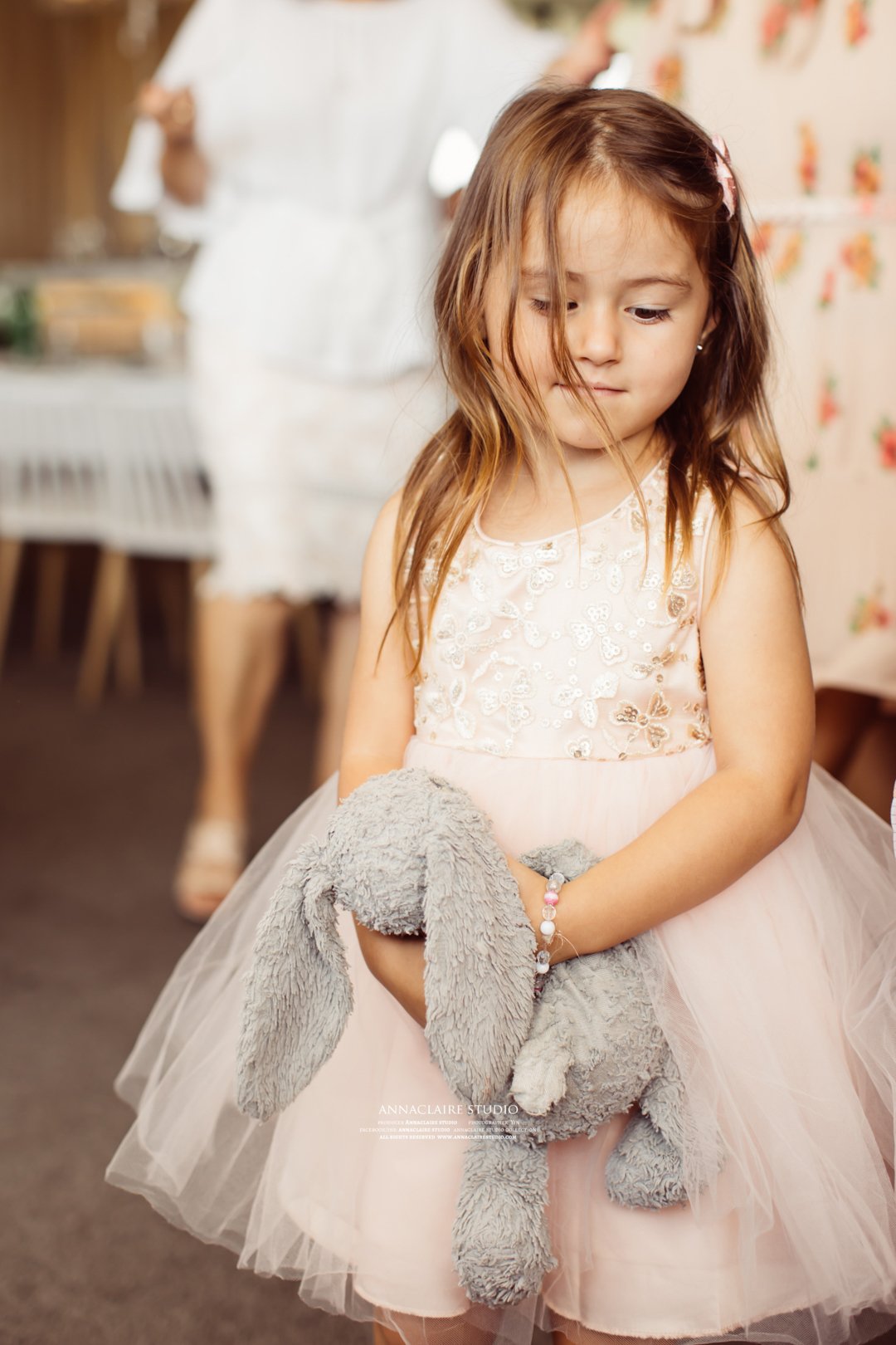 A young girl in a light pink dress with floral embroidery holds a gray stuffed bunny. She has long, wavy brown hair and a pink hair clip, and is wearing a bracelet with beads. She appears to be at a social event or celebration, with people and decora