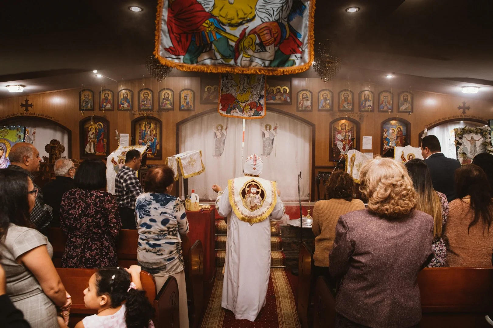 People attending a religious service inside a church, facing the altar where a priest dressed in white robes with embroidered symbols stands with his back to the congregation. The church interior features wooden paneling, icons, and religious paintin