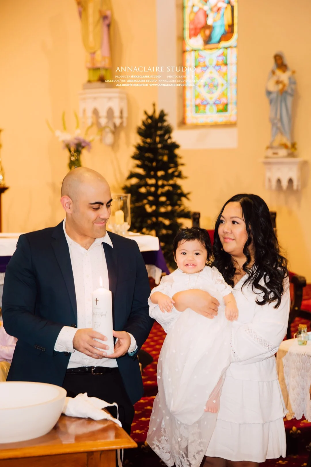 A family at a baptism or christening ceremony inside a church. The man is holding a lit candle with a cross and text on it. The woman is holding a smiling baby dressed in white. The background features a Christmas tree, stained glass window, and reli