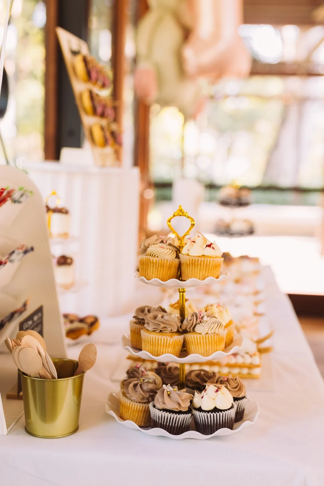 A three-tiered cupcake stand displaying assorted cupcakes with swirled frosting and decorative toppings, set on a white tablecloth at a bakery or event preparation area.