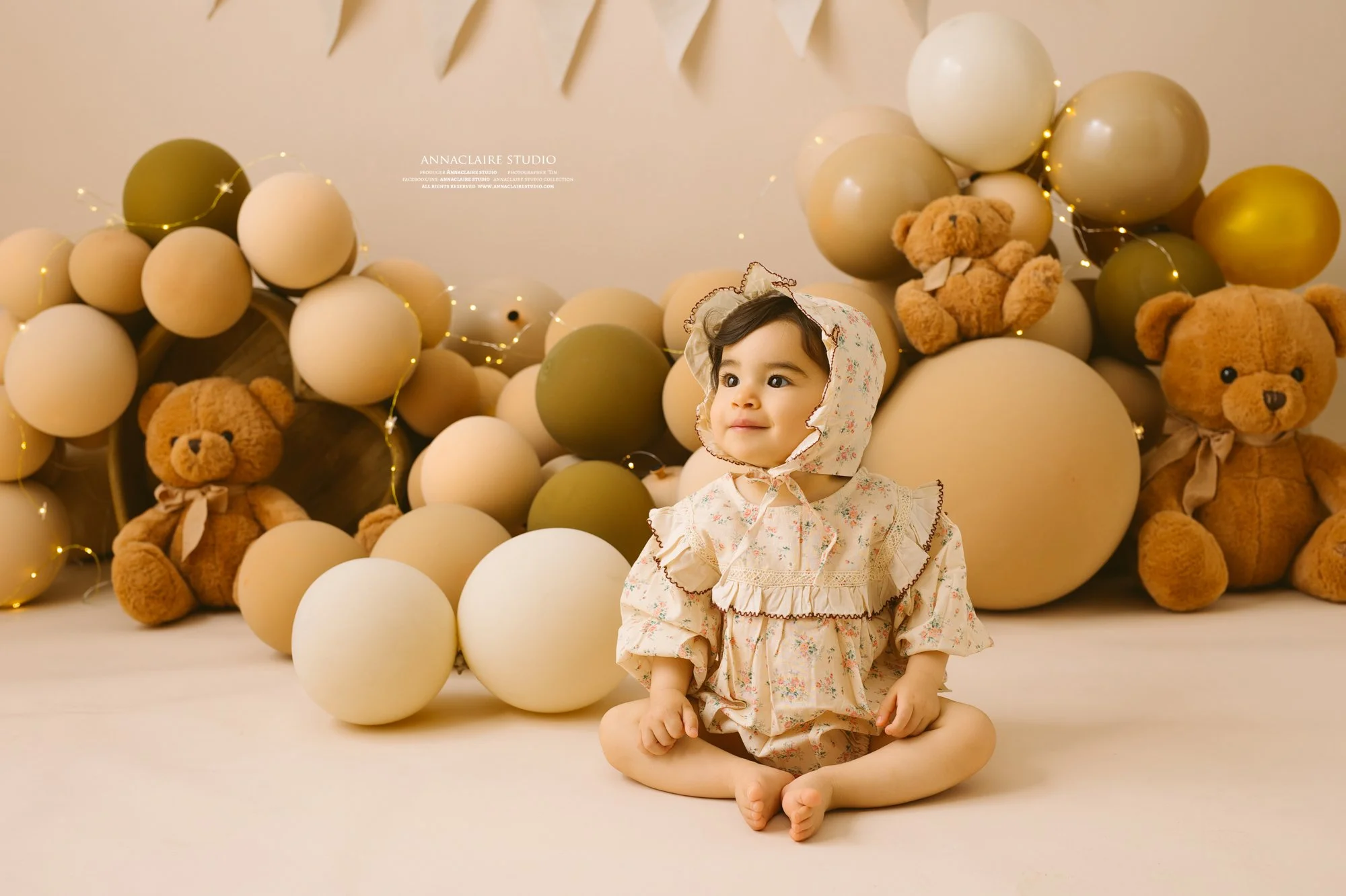 A young girl sitting on the floor surrounded by beige, cream, and green balloons along with teddy bears, with soft lighting creating a warm and cozy atmosphere.
