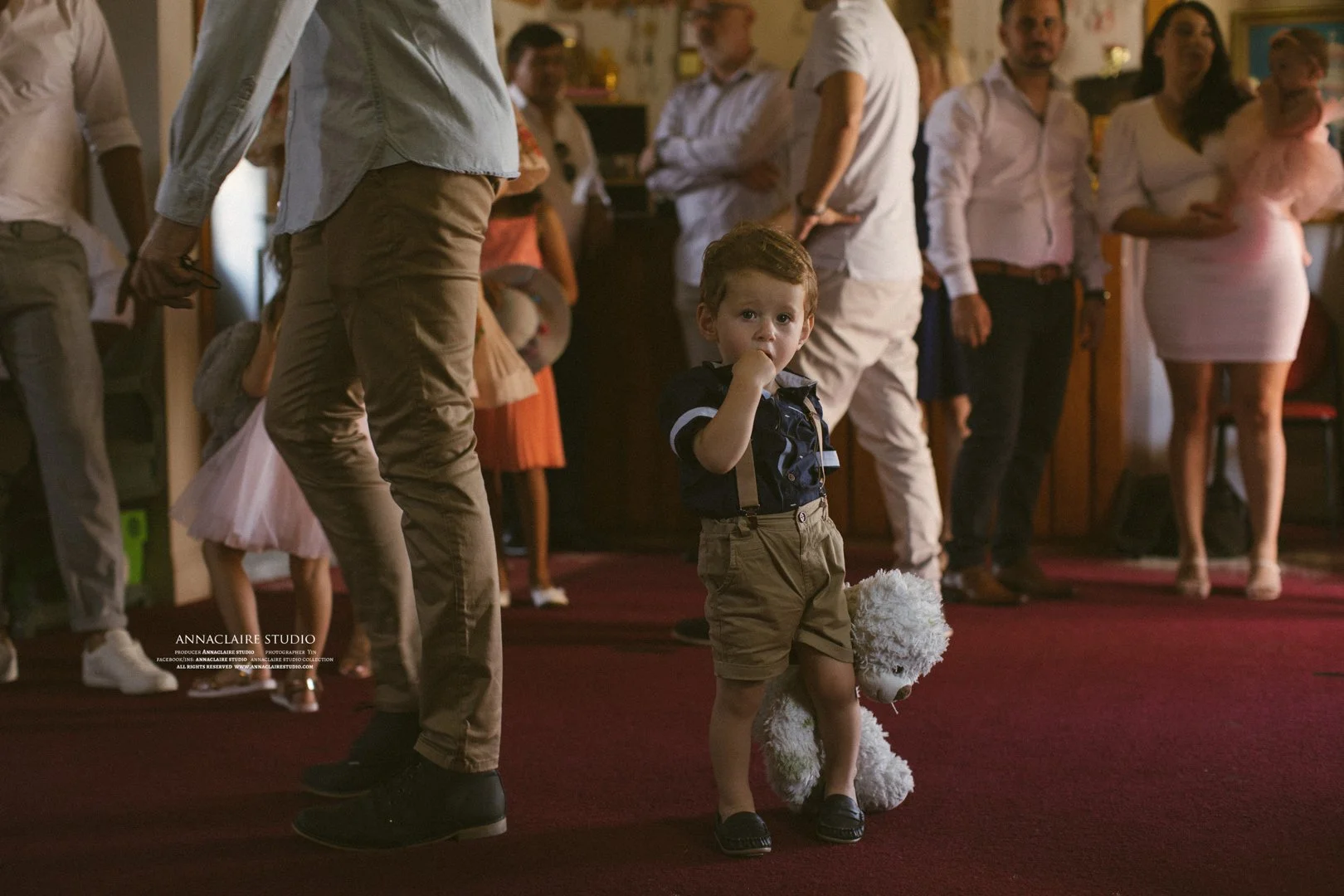 A young boy with brown hair in suspenders and shorts standing on a red carpet holding a stuffed white teddy bear, with a group of adults and children in the background
