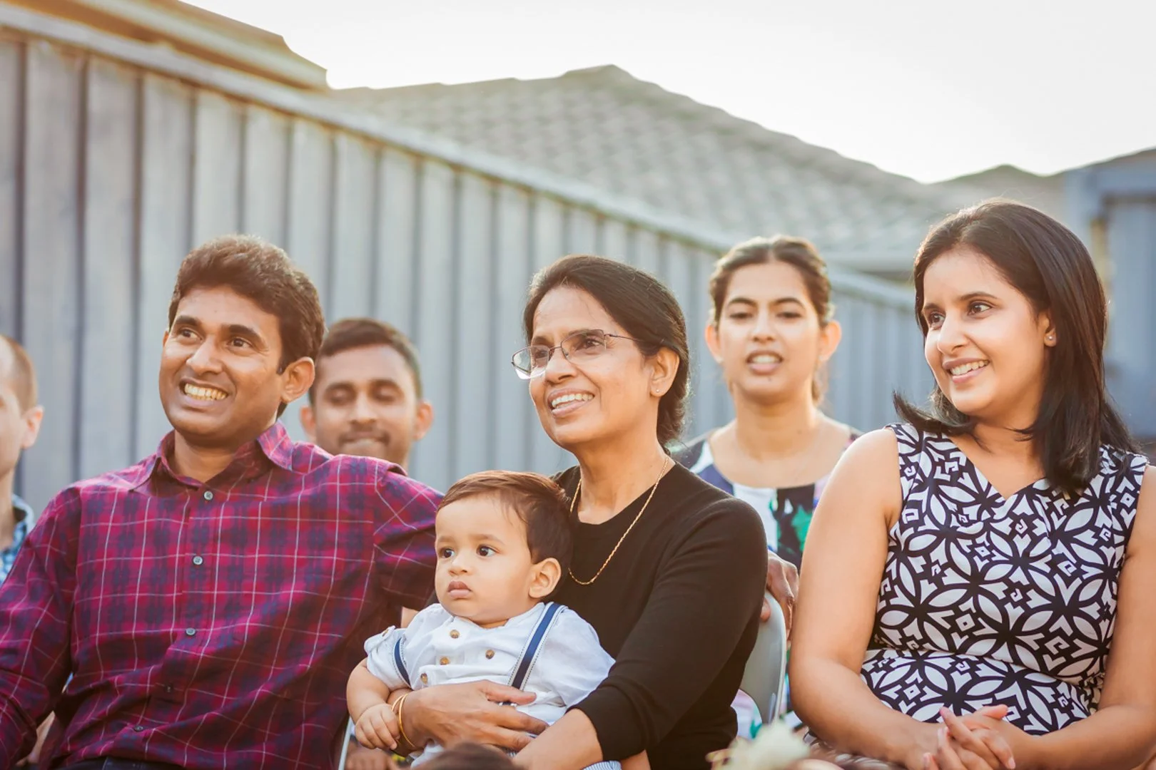 Group of people outdoors, smiling and sitting together at an event, with a woman holding a young boy on her lap.