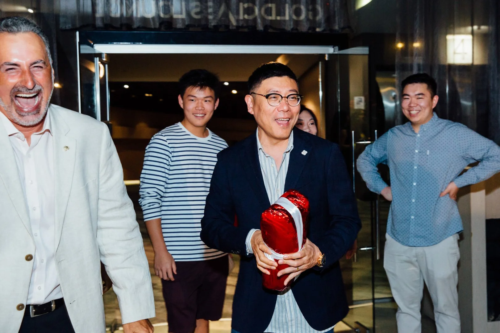 A group of five people smiling, with one man holding a red gift wrapped in a shiny red cloth and white ribbon.