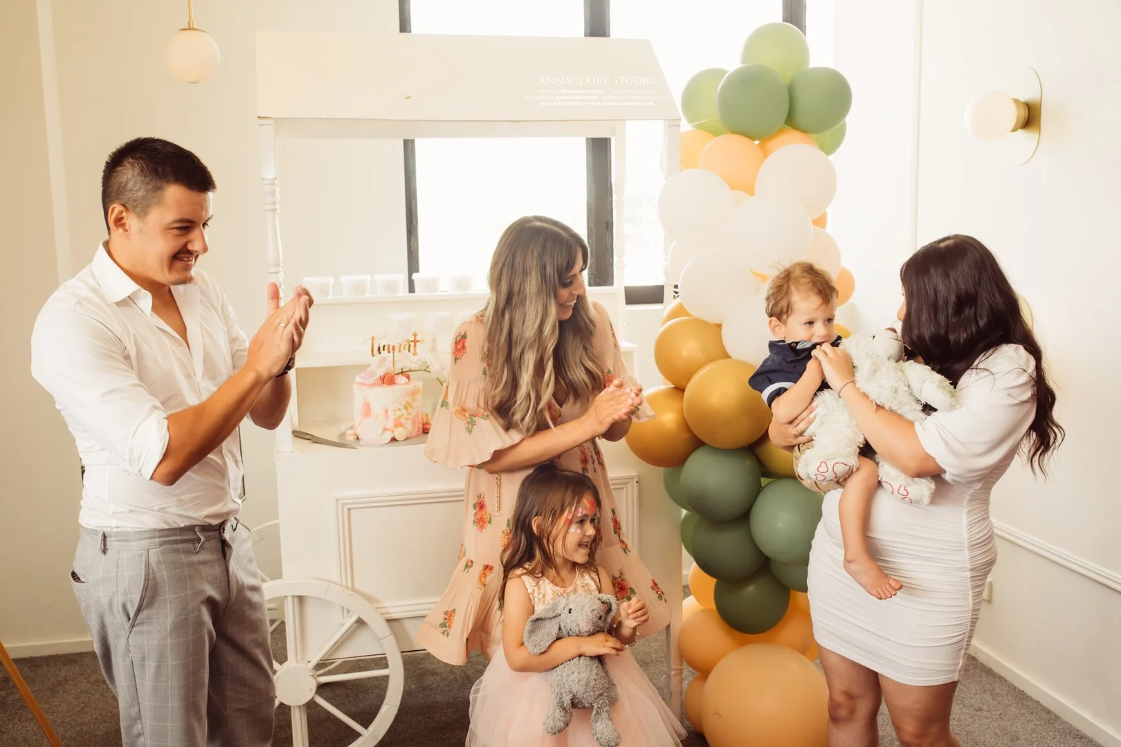 A group of people celebrating a child's birthday indoors, with balloons and a decorated cake in the background. A woman is holding a young girl with a stuffed animal, while another woman and a man are clapping and smiling.