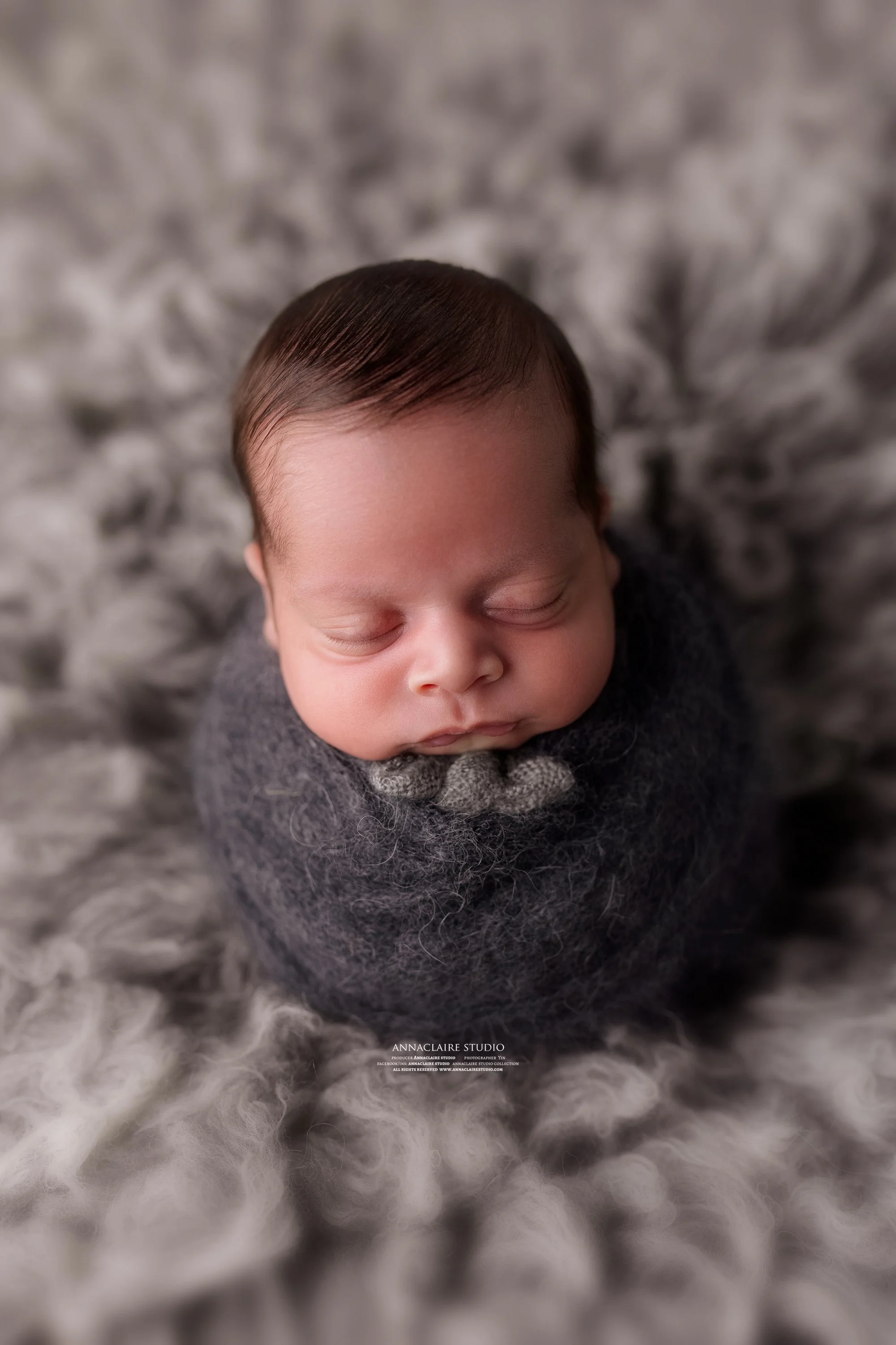 A sleeping newborn baby wrapped in a dark gray blanket, lying on a soft, fluffy gray and white fur blanket.