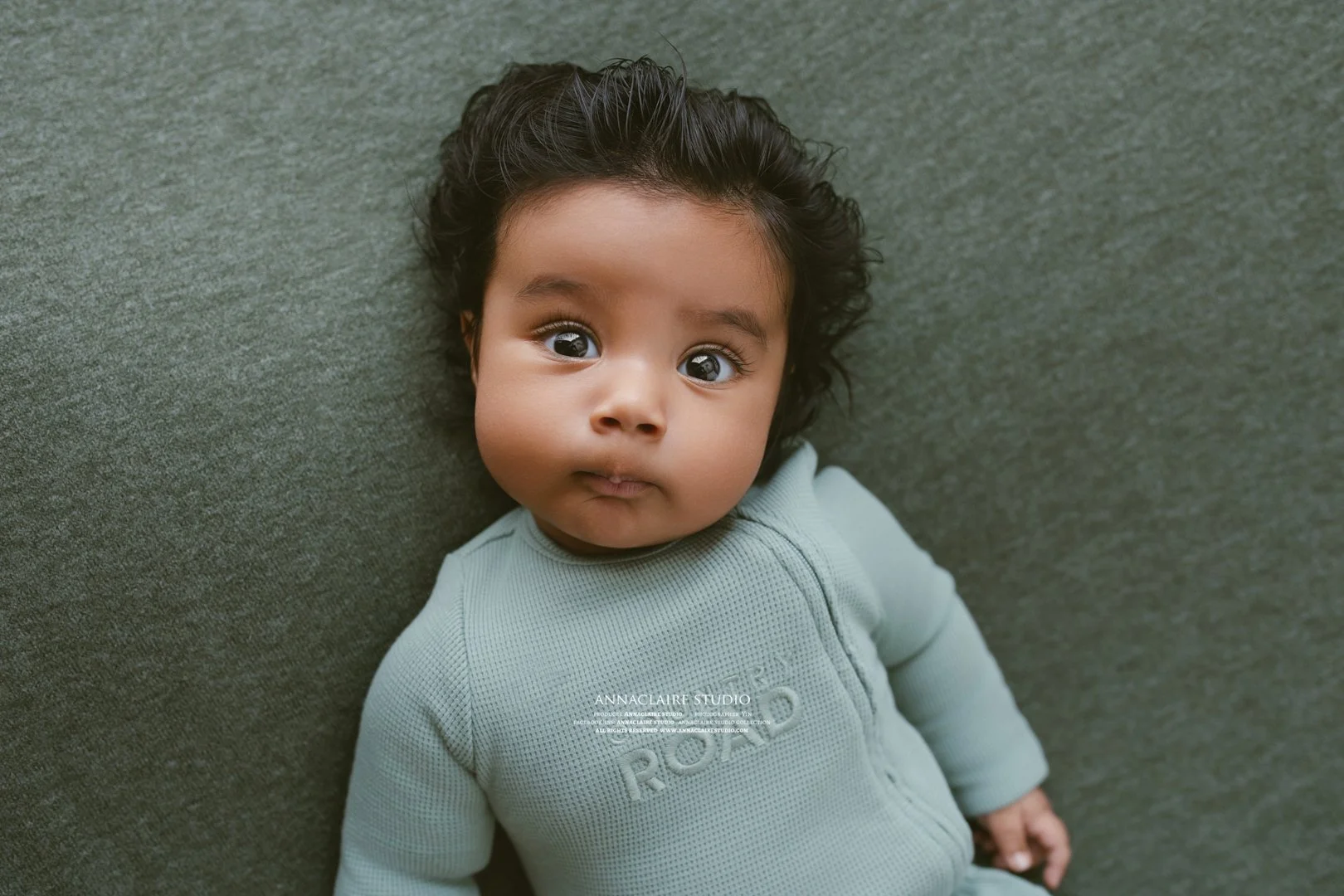 Close-up of a young child with dark hair and big eyes lying on a gray surface, wearing a light green shirt.