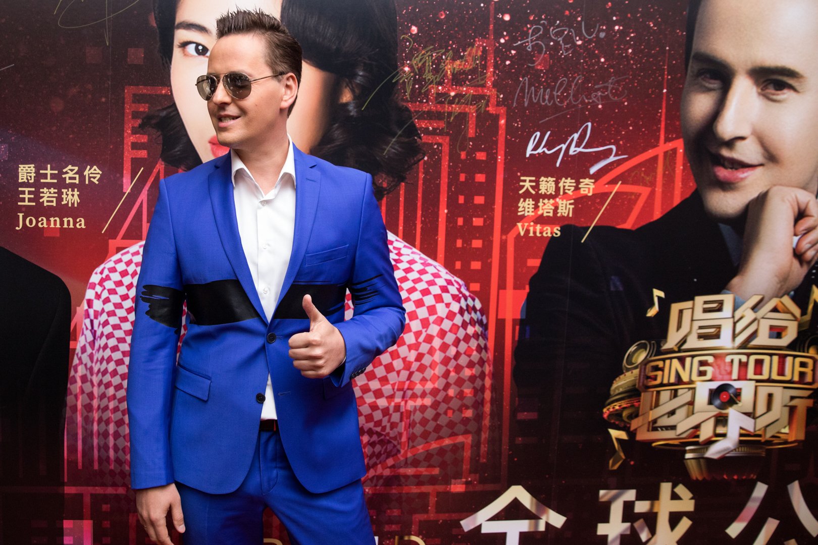 Man in blue suit and sunglasses giving a thumbs up standing in front of a red background with Chinese characters, signatures, and images of two women.