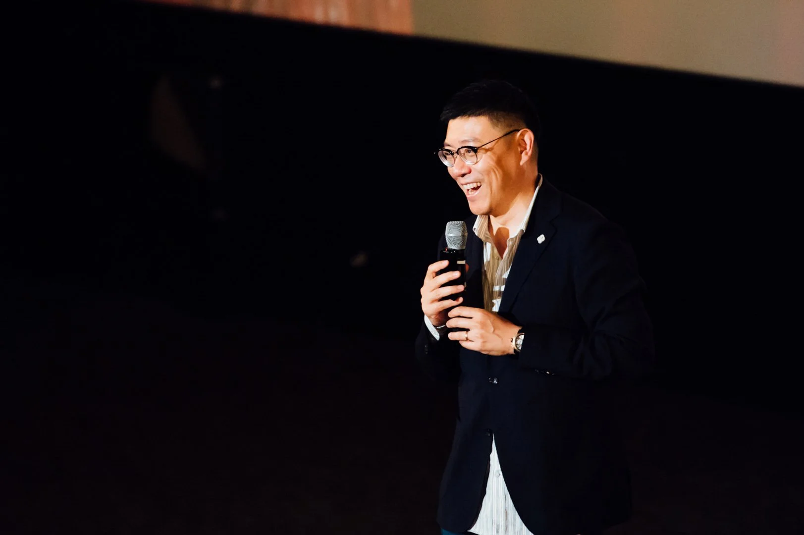 Man in black blazer and glasses speaking into a microphone, smiling, against a dark background.