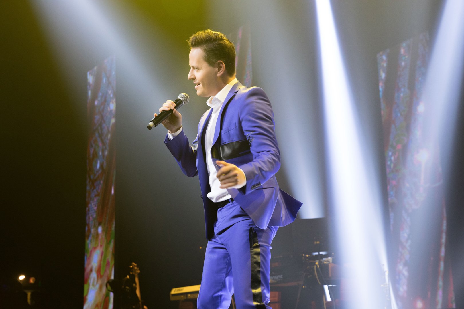A male singer in a blue suit performing on stage with a microphone, illuminated by stage lights.