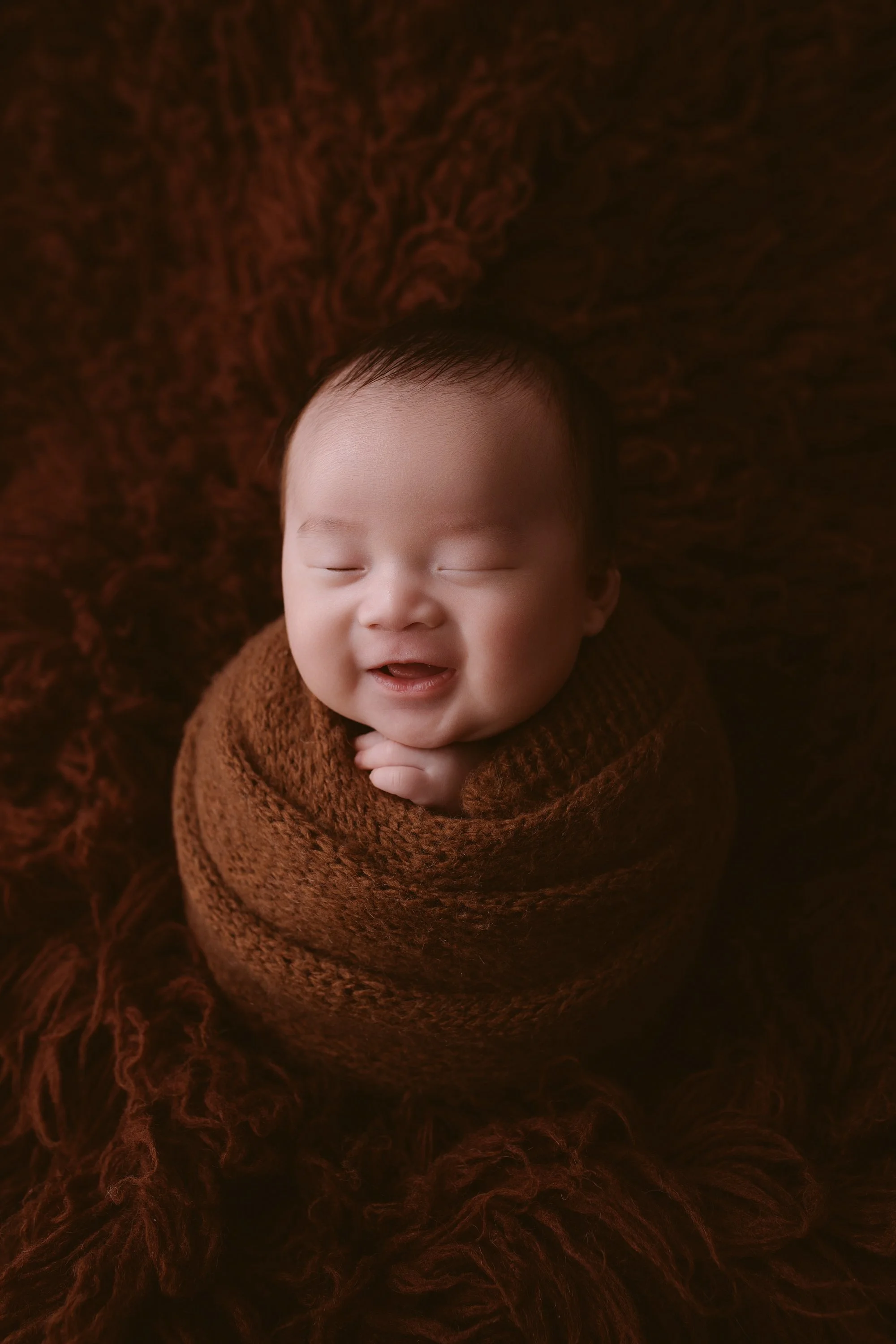 Smiling baby wrapped in a brown blanket with a matching brown fluffy background.