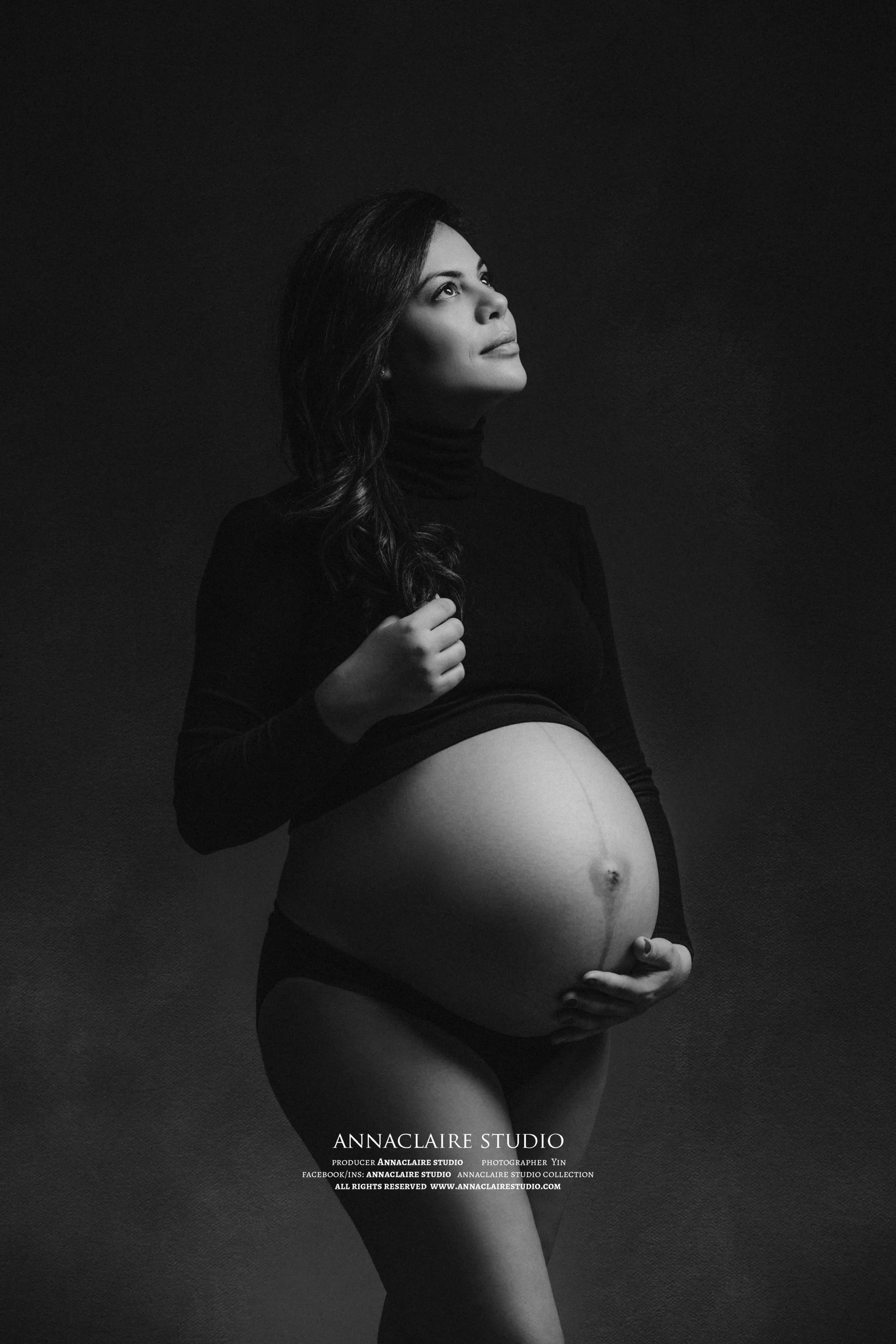 Black and white portrait of a pregnant woman in a black long sleeve shirt, looking up, holding her belly.