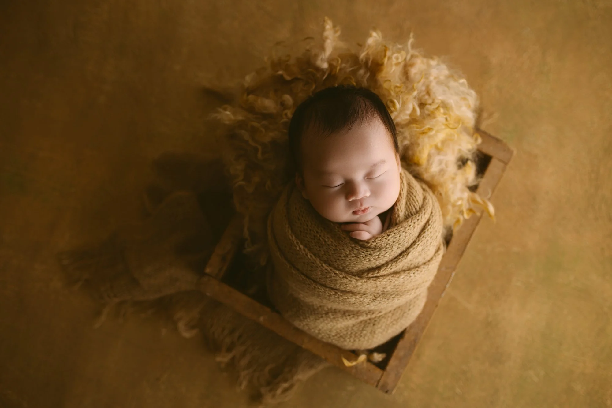 A baby wrapped in a beige blanket, sleeping with hands under chin, on a furry yellow rug, inside a small wooden box, on a yellowish floor.