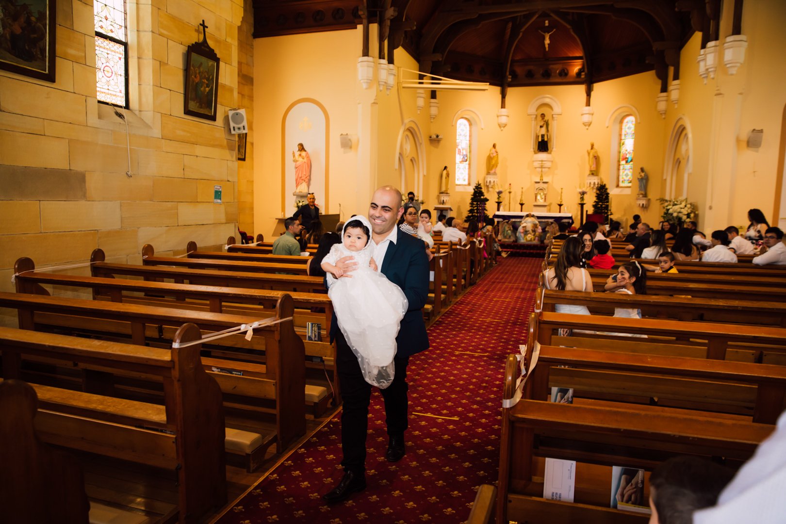 A man in a dark suit holding a young girl in a white dress inside a church during a religious ceremony.
