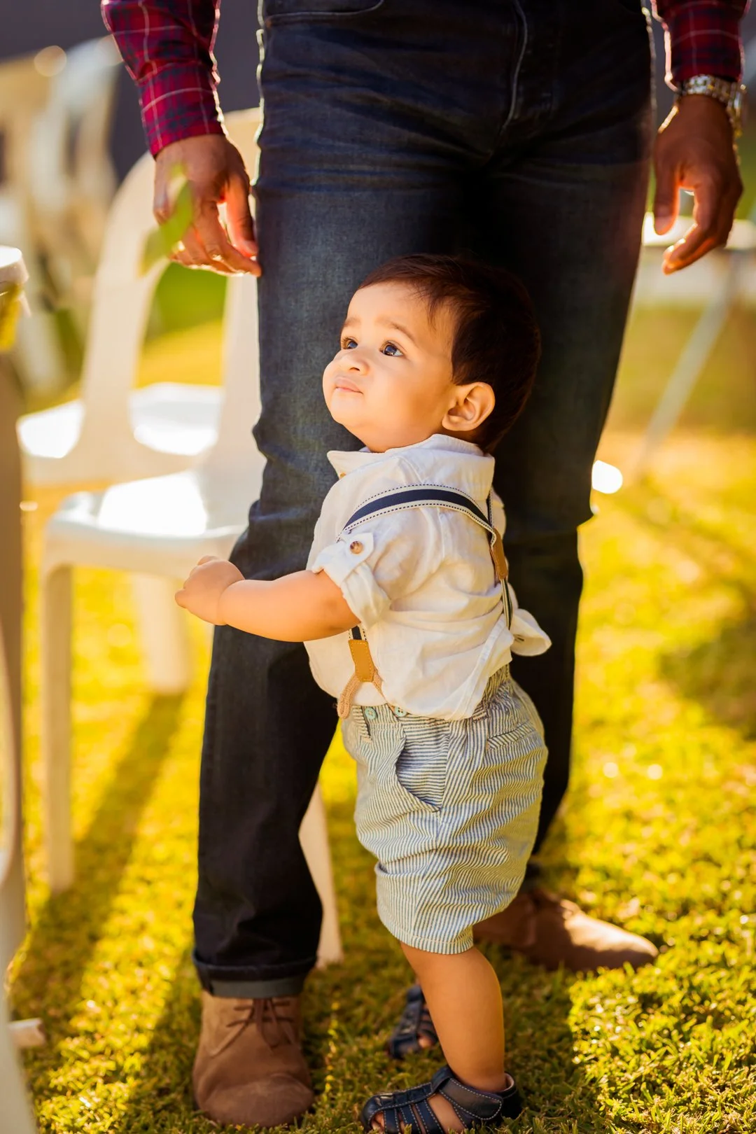 A young boy holding onto an adult's leg outdoors, looking upward with a curious expression, surrounded by chairs and warm sunlight.