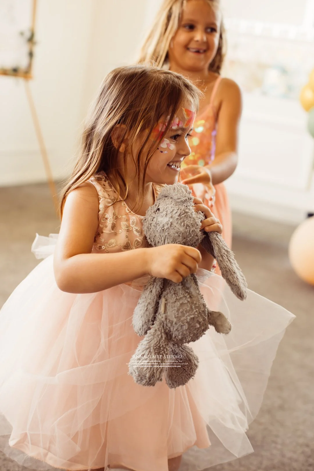 Two young girls, one in a peach tulle dress, playing with a stuffed gray bunny at a party with balloons and decorations.