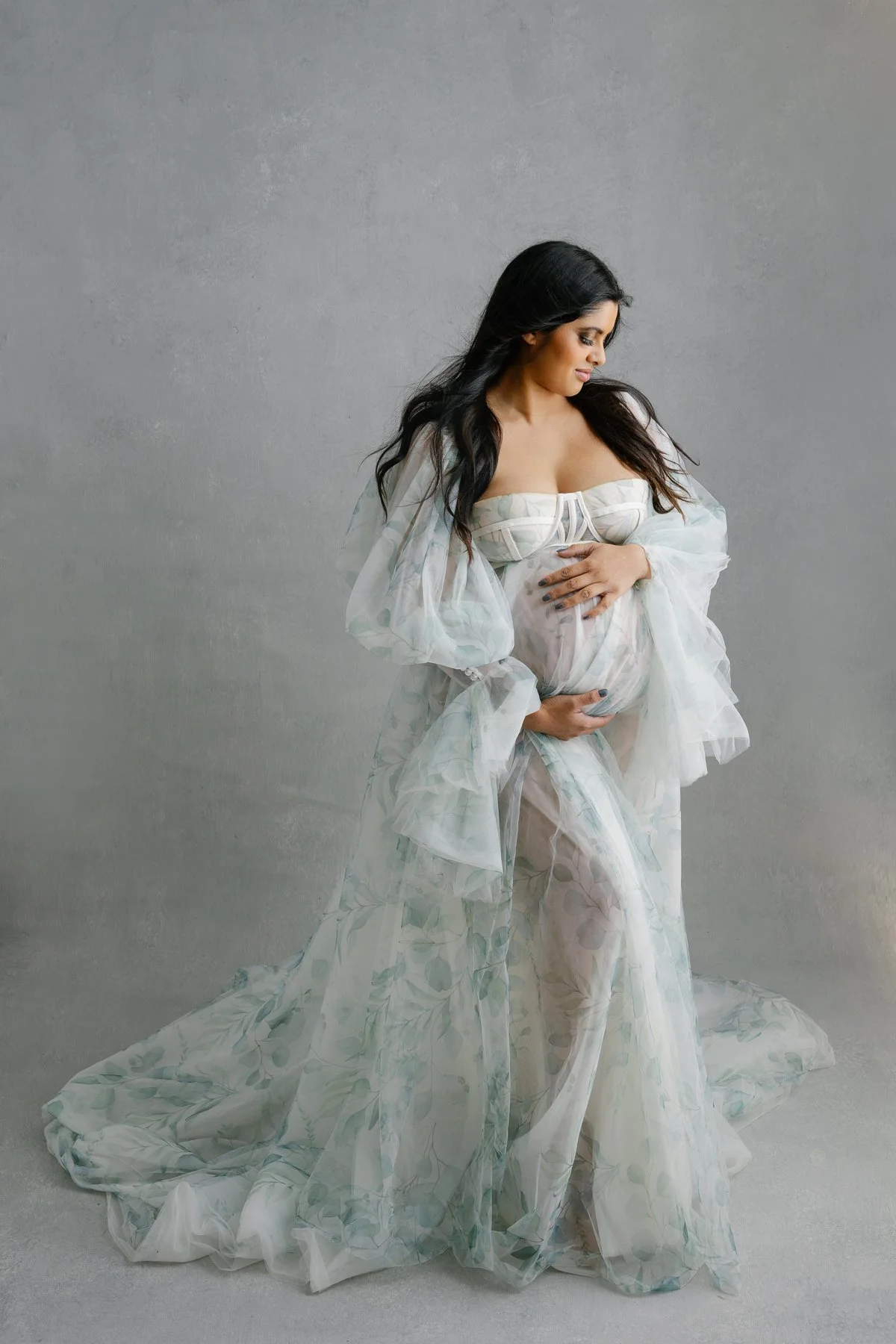 Pregnant woman in a sheer, flowing, pastel-colored dress posing gently with her hands on her belly against a neutral background.