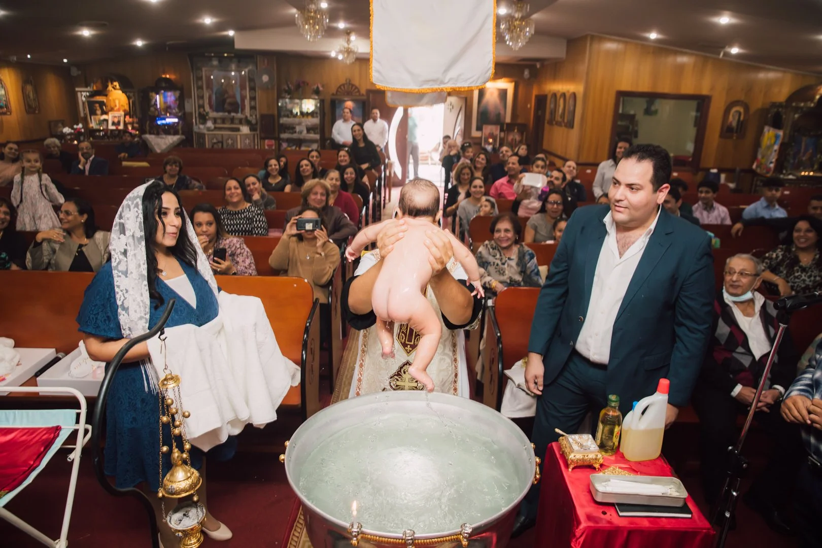 A priest performs a baptism ceremony in a church, holding a naked baby over a baptismal font filled with water as a crowd of family and friends look on, some smiling and taking photos.
