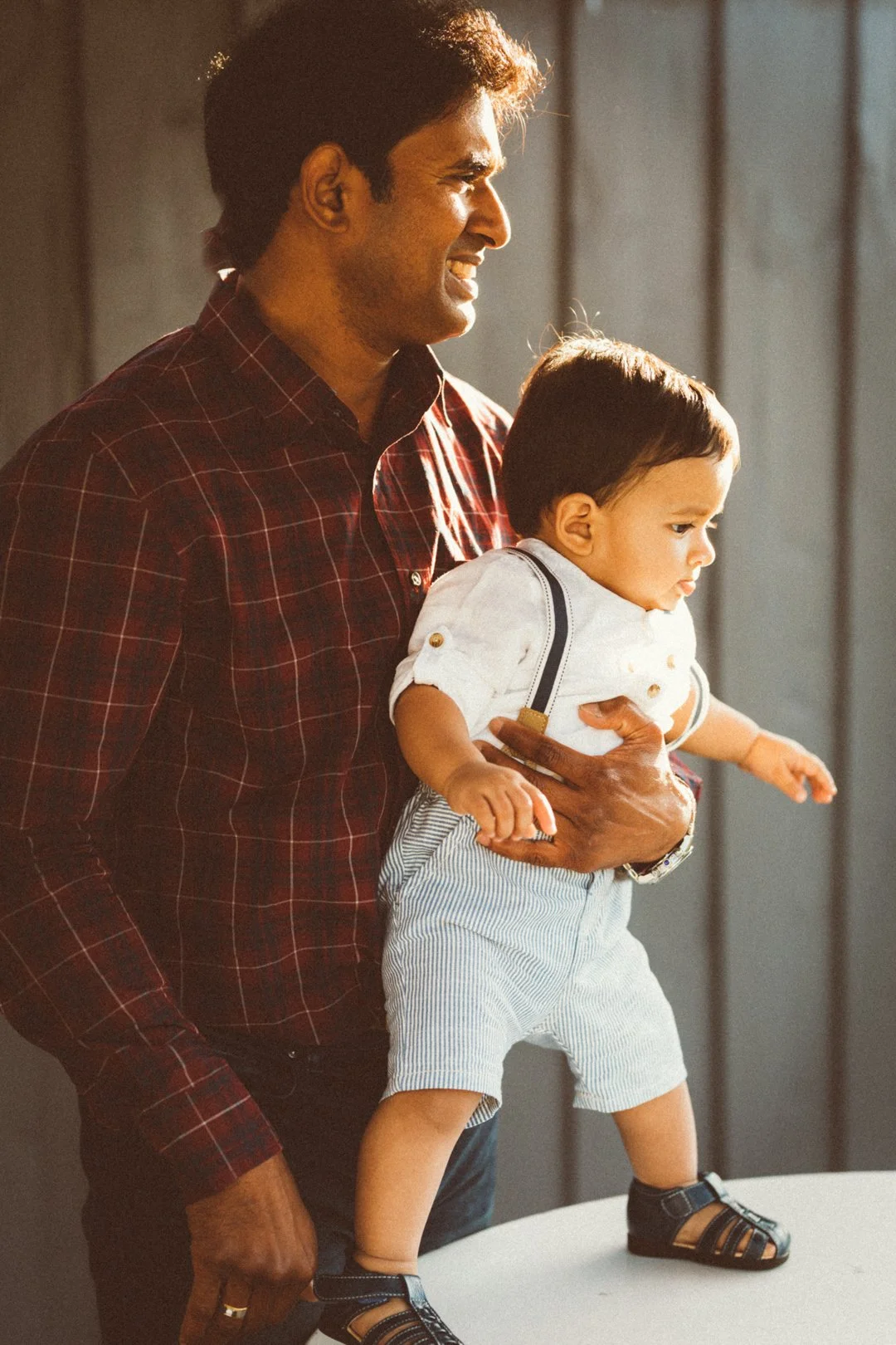 A man in a red plaid shirt holding a young child standing on a table, with both looking to the side. The man smiles while the child appears focused.