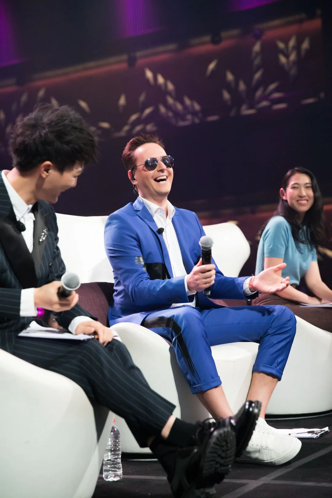 Three people sitting on a stage, one in the center wearing a blue suit and sunglasses, laughing with a microphone, while the others hold microphones and look on, with a dark background and purple lighting.