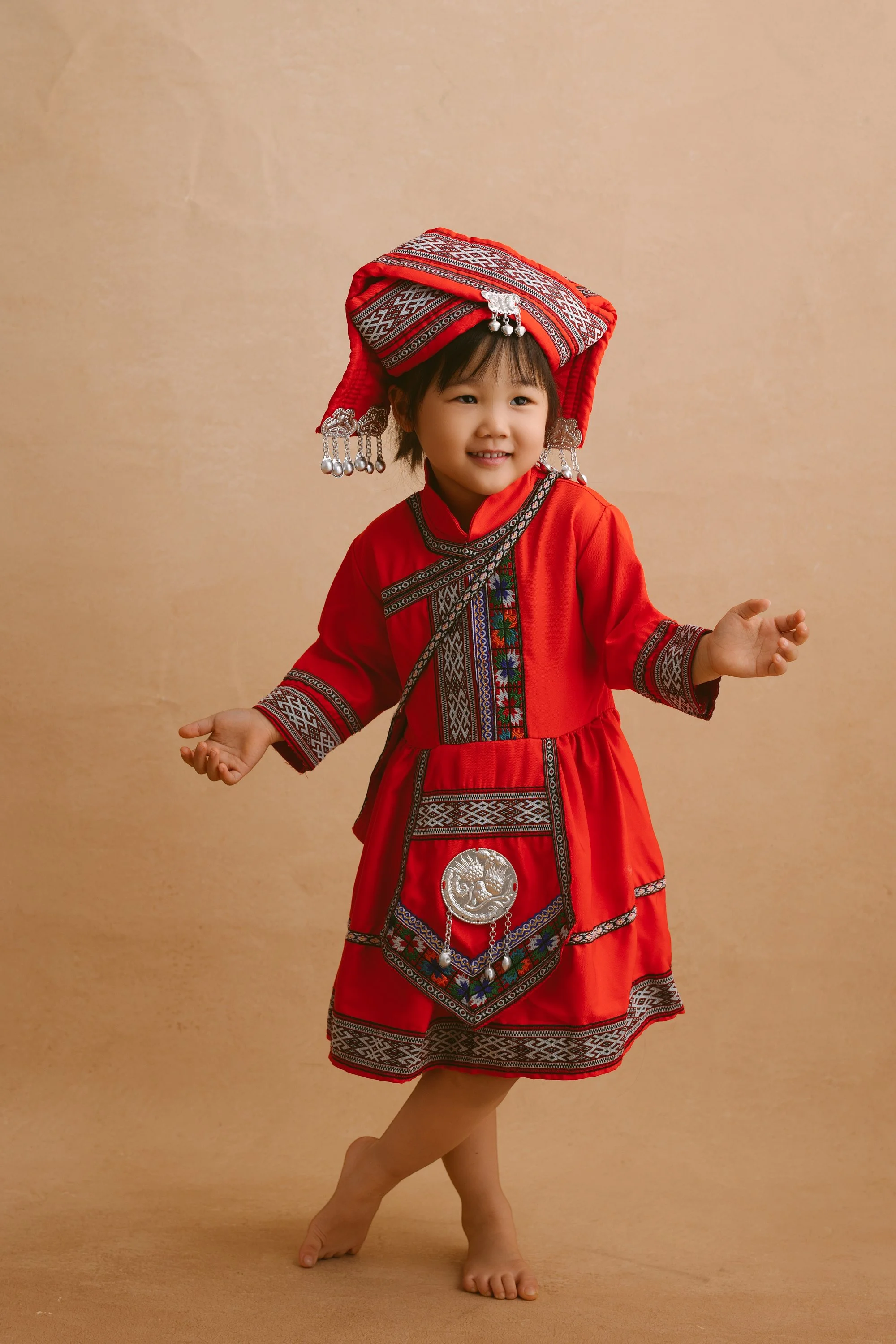 Young girl dressed in traditional red ethnic attire with intricate silver and embroidery details, smiling and posing barefoot against a beige background.