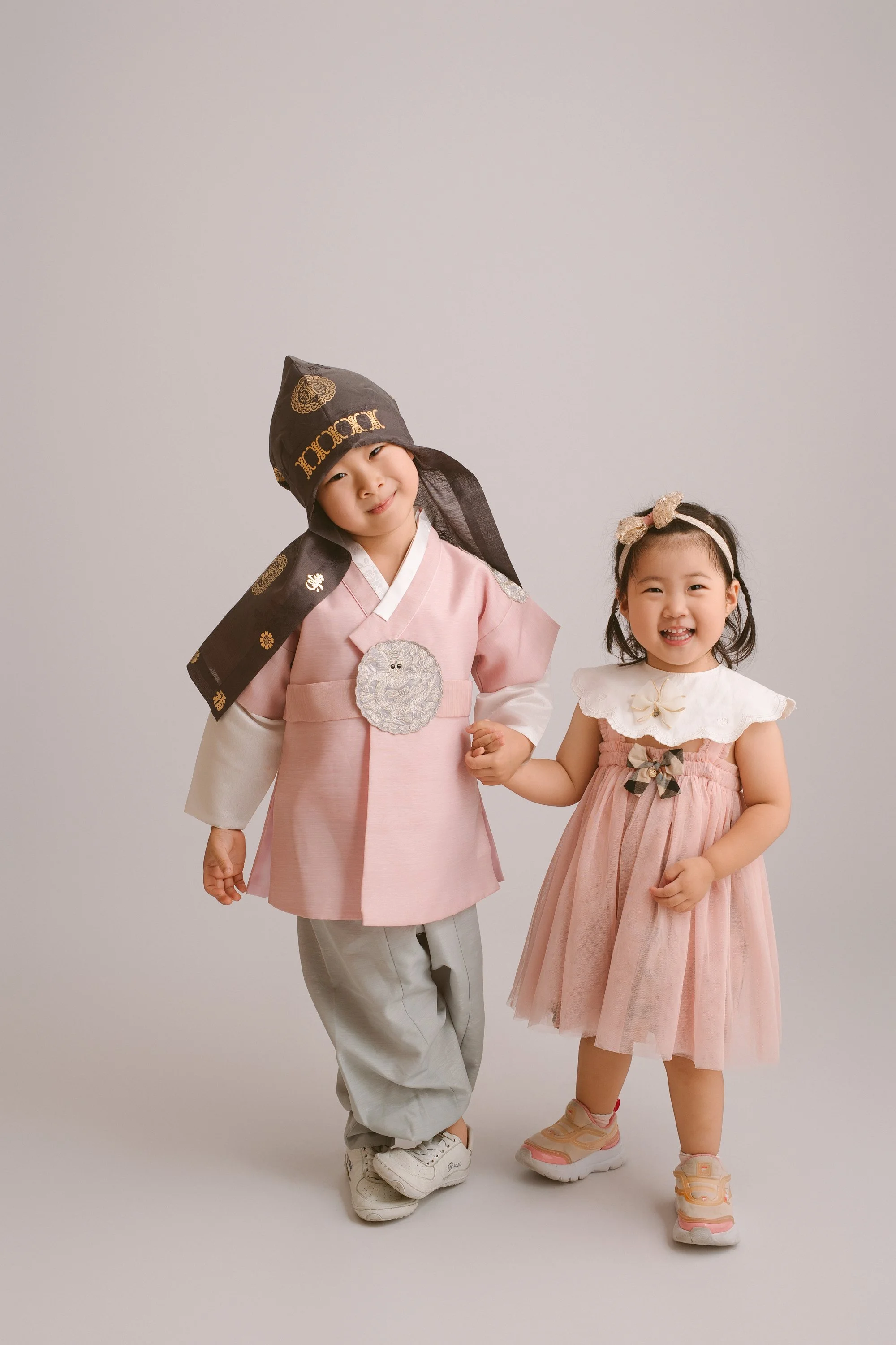 Two young children, a boy and a girl, holding hands and smiling at the camera. The boy is dressed in traditional Korean hanbok with a pink top, grey pants, and a black hat. The girl is wearing a pink dress with a white collar and a bow, with a headba