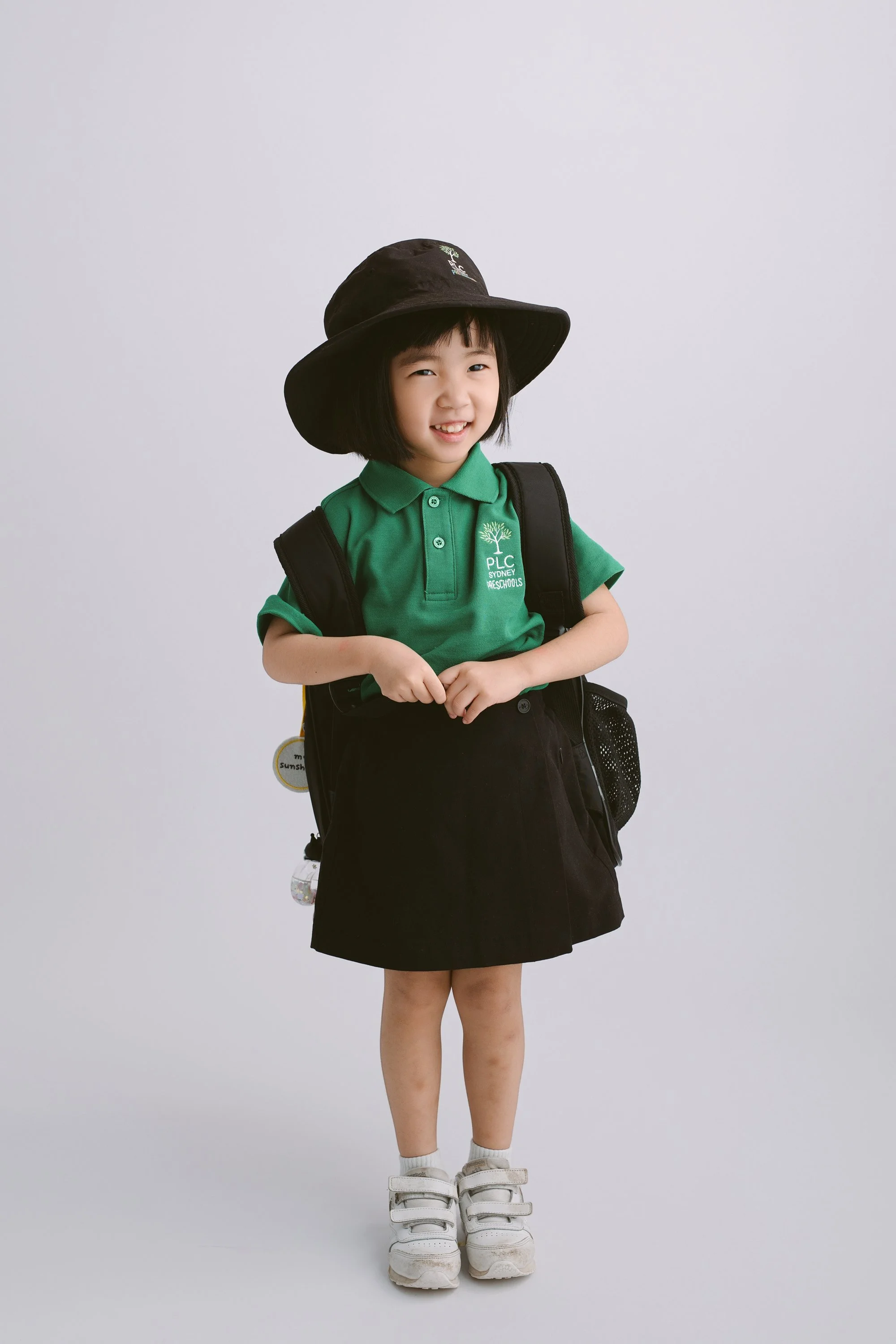 A young girl in a green school uniform, black skirt, white sneakers, and a large black hat, smiling and standing against a plain white background.