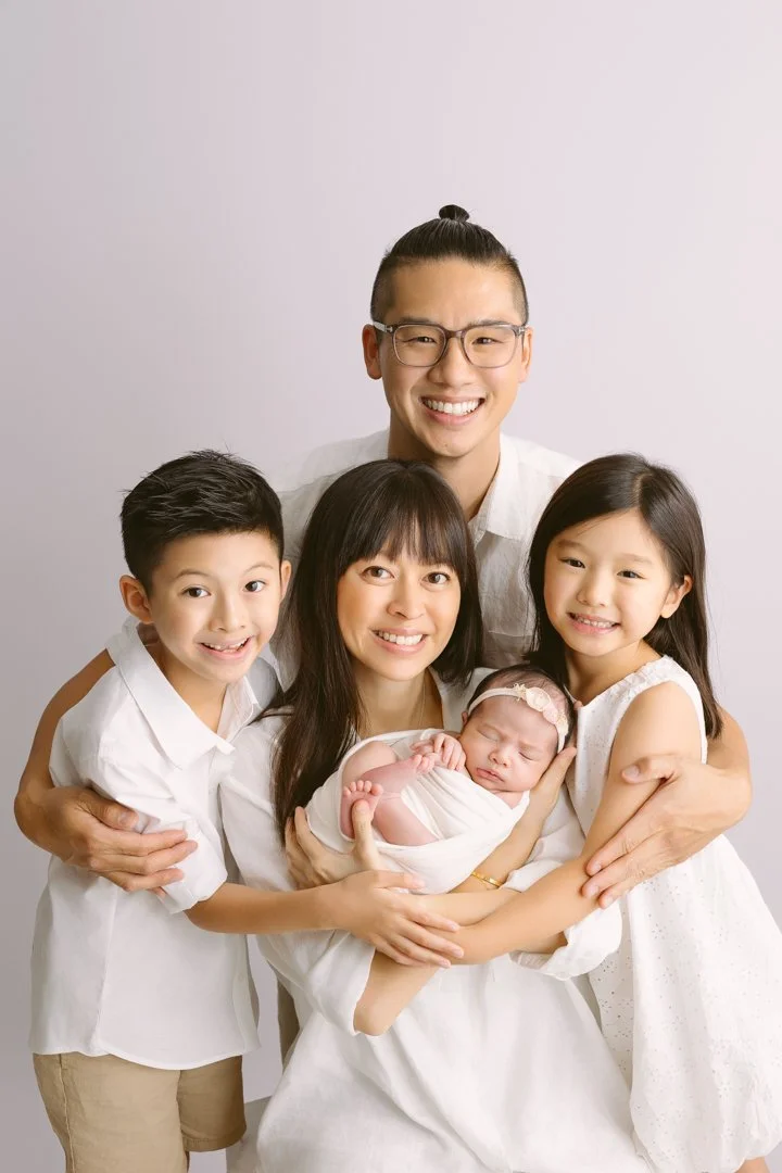 Family of five, including a newborn baby girl, posing together smiling against a plain background.
