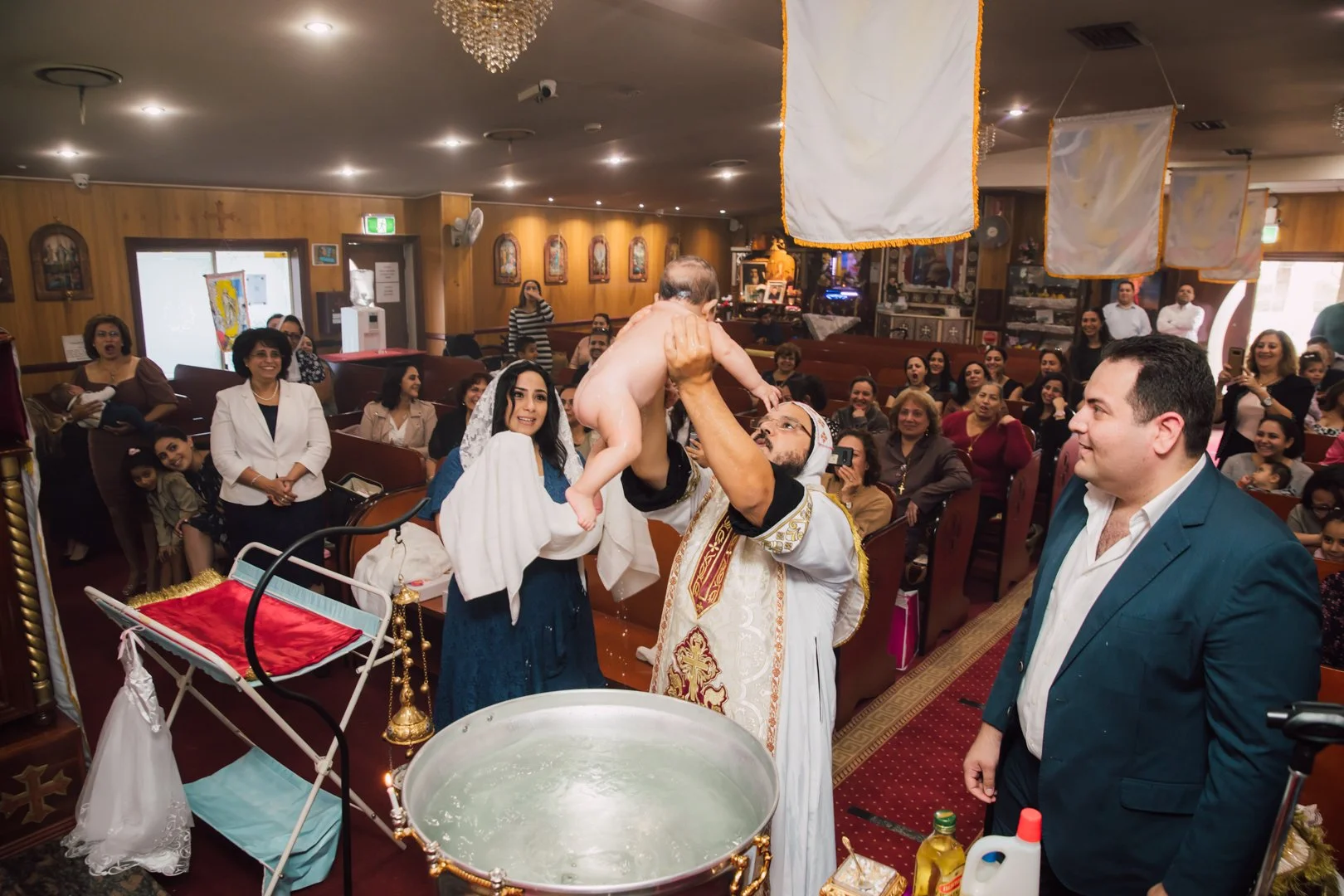 A priest lifts a naked baby during a baptism ceremony in a church filled with smiling guests.