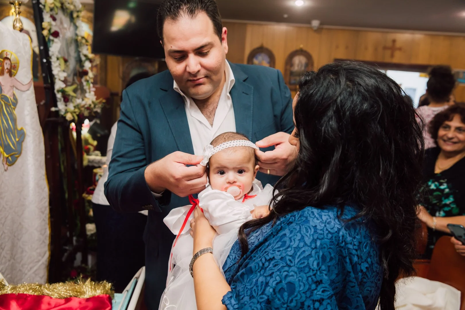 A man dressed in a suit is helping a woman dress a baby for a religious ceremony. The baby has a pacifier and is wearing a white outfit with red ribbons. The woman has long black hair and is wearing a blue patterned dress. They are inside a decorated