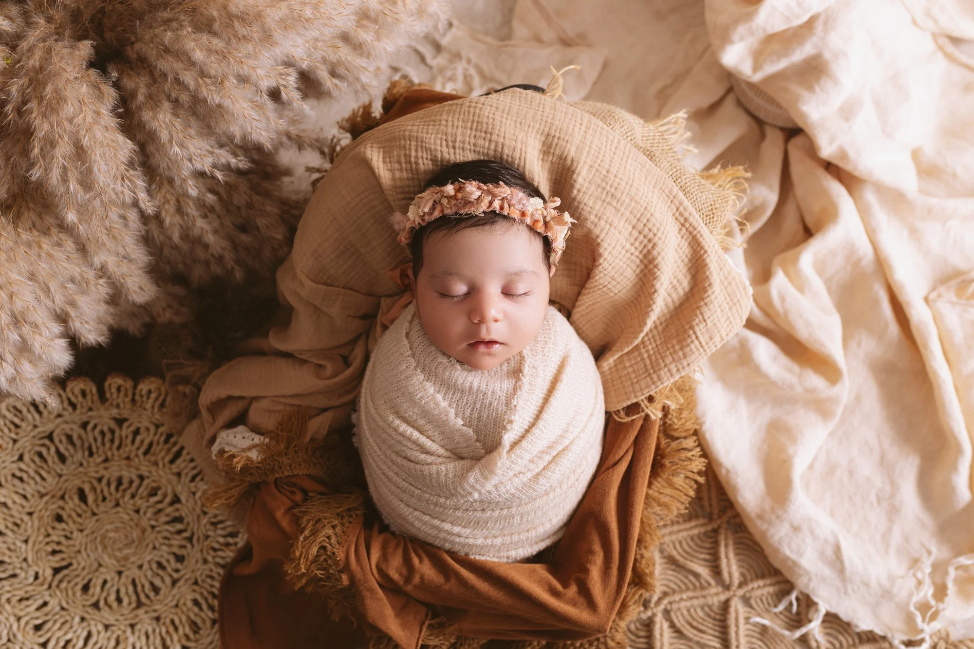 A sleeping baby wrapped in cream-colored swaddle, wearing a floral headband, lying on a cozy blanket surrounded by pampas grass and textured fabrics.
