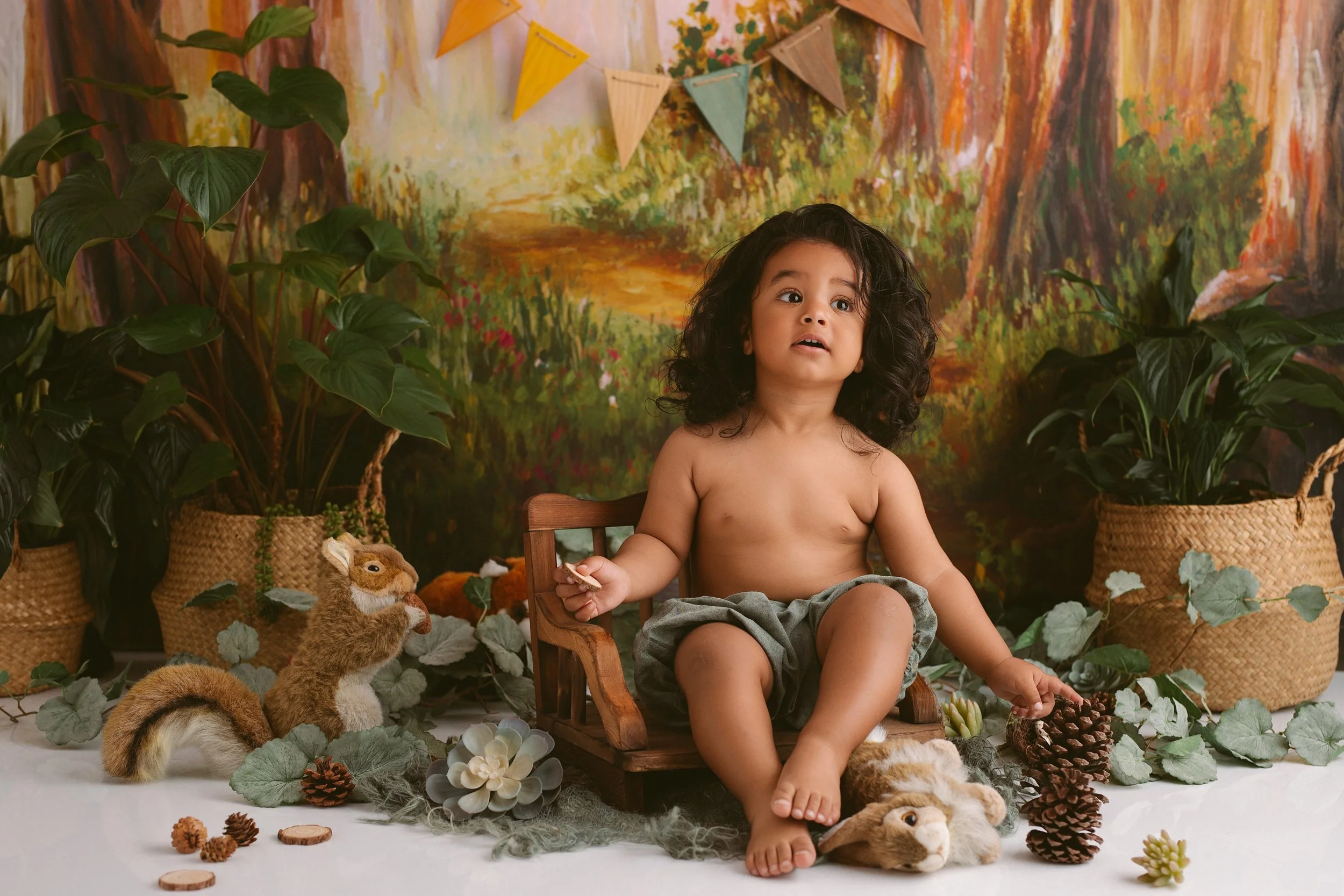 A young child with curly hair, shirtless and wearing gray shorts, sitting on a wooden chair in a nature-themed setting with plants, pinecones, and stuffed animals, against a painted forest background.