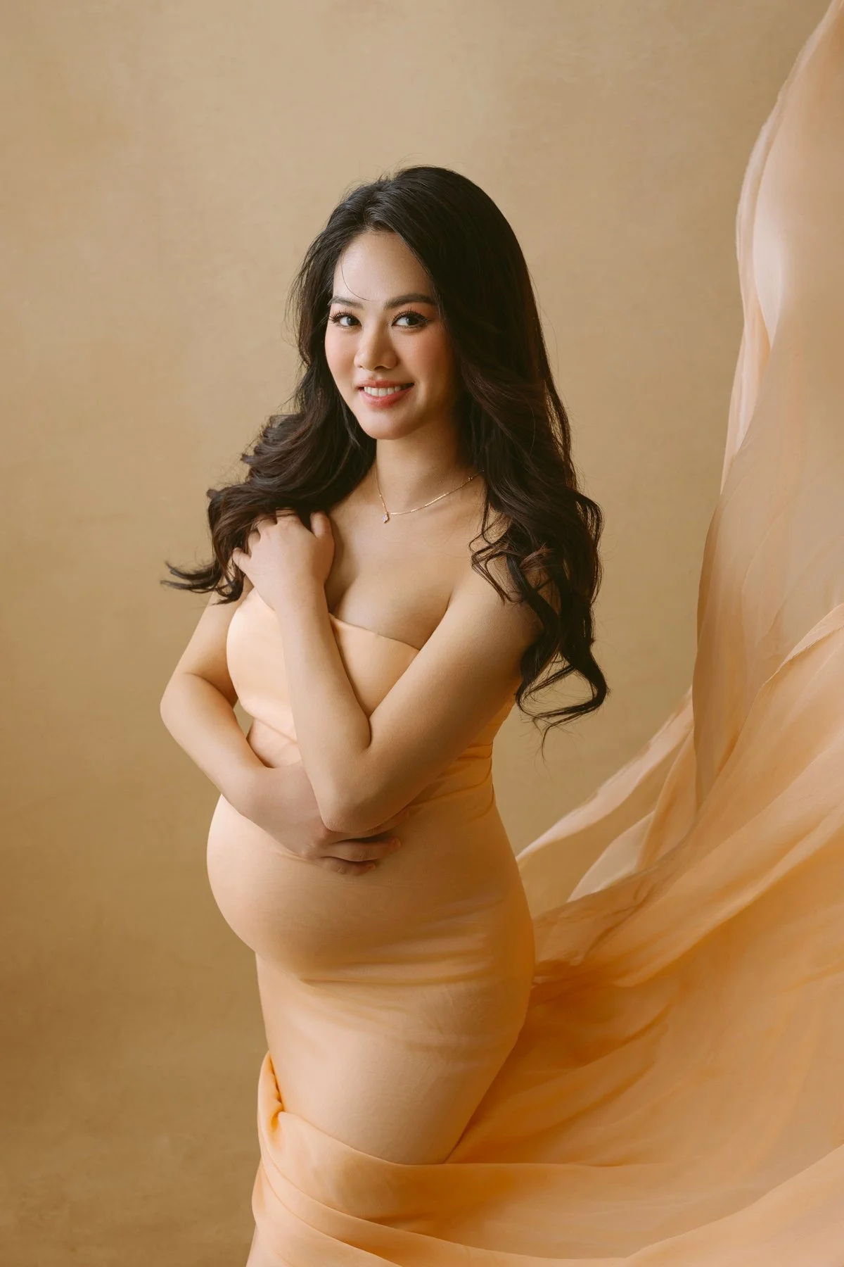A smiling woman with long, dark hair wearing a nude-colored dress with a flowing train, standing against a beige background.