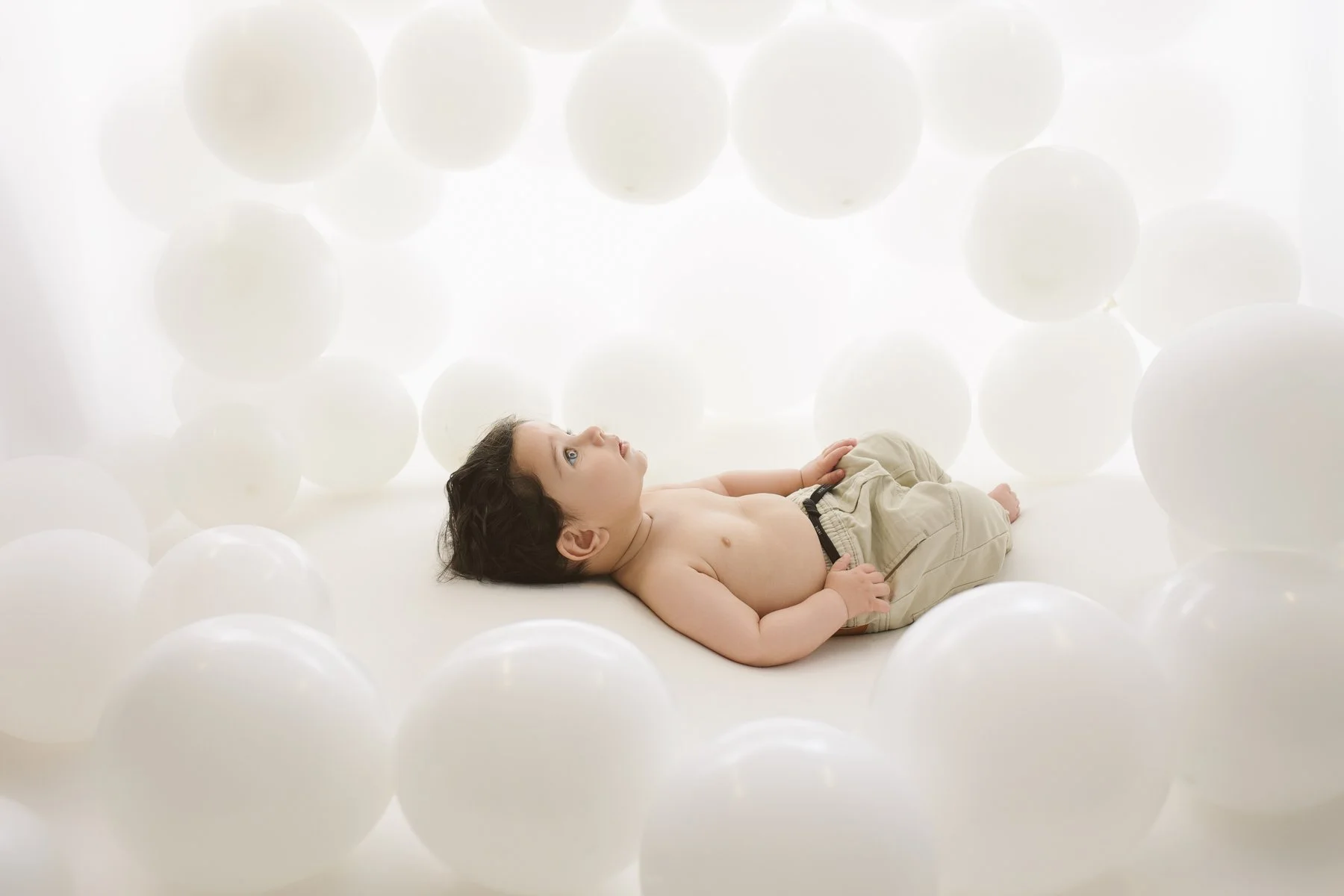 A young boy lying on a white surface, surrounded by large white spherical objects, looking upward with a curious expression.