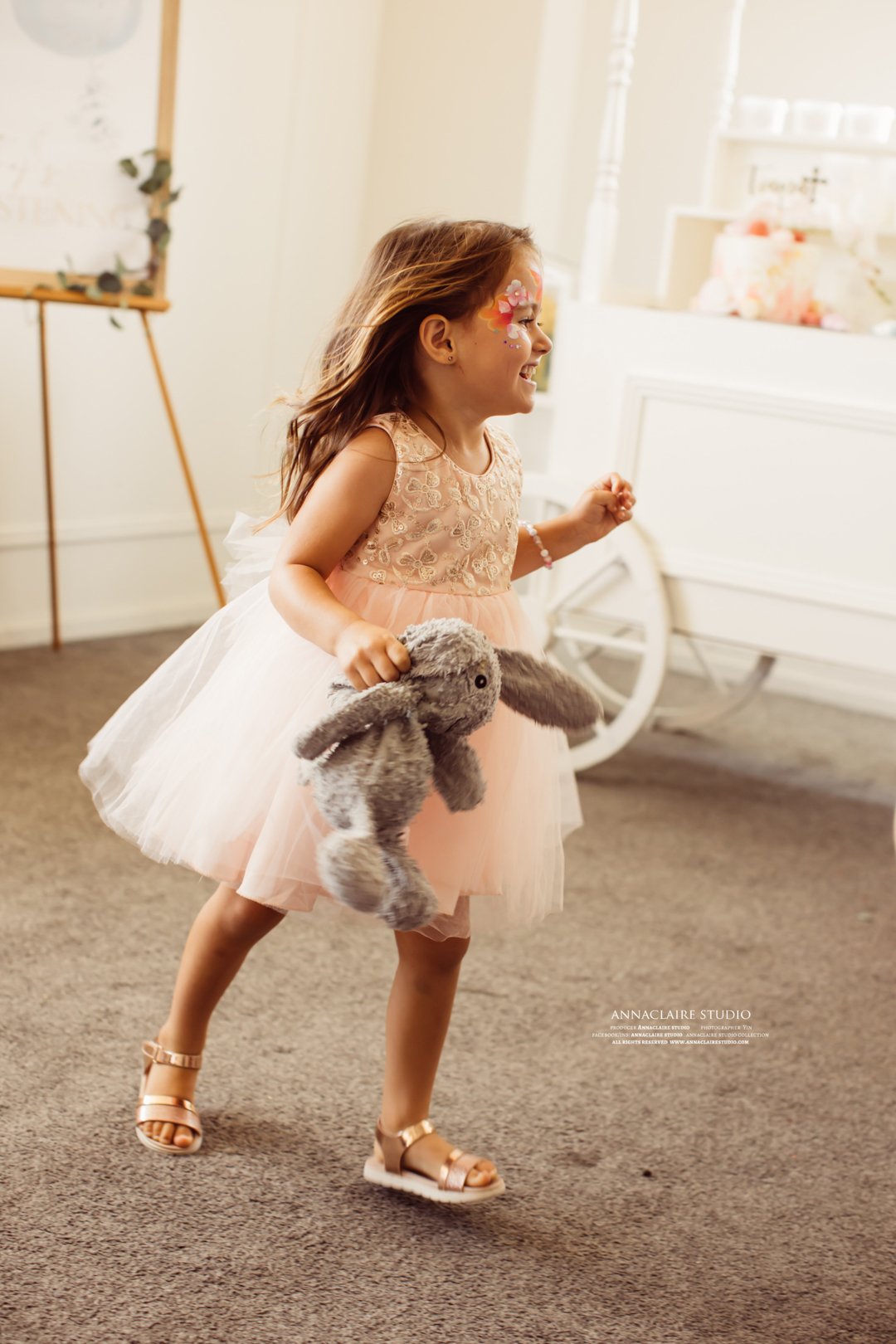 A young girl with face paint and a pink dress running with a stuffed bunny toy inside a bright, decorated room.