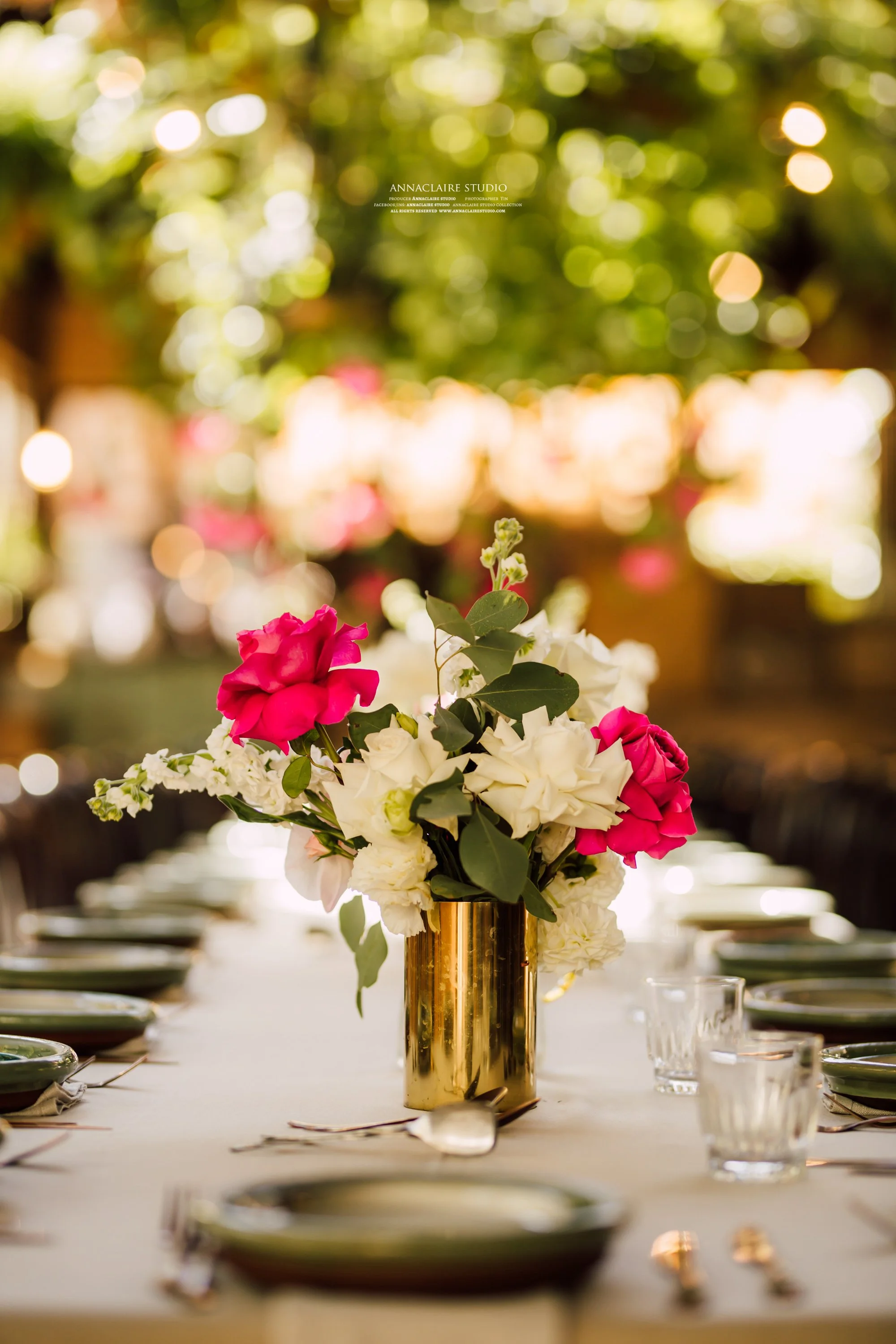 A table decorated with a floral centerpiece of pink and white flowers in a gold vase, set with plates, glasses, and cutlery, in an outdoor setting with blurred background and trees.