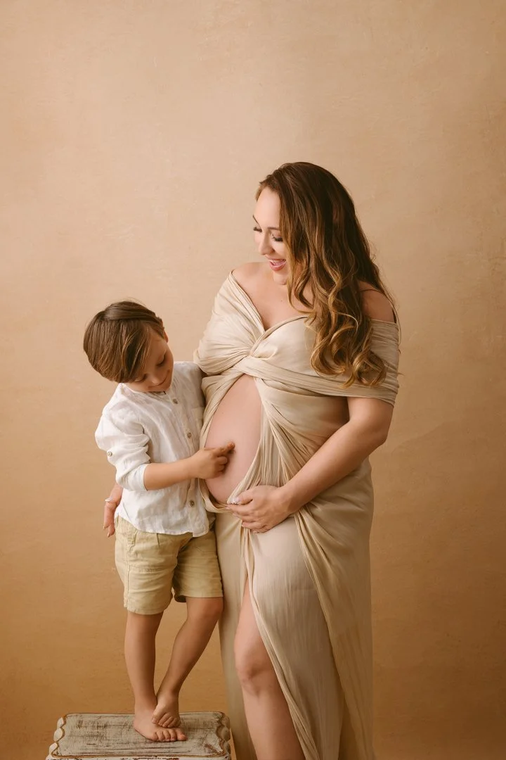 A pregnant woman in a beige dress smiling as she touches her belly, standing next to a young boy who is gently touching her belly and looking down. They are indoors against a plain beige wall.