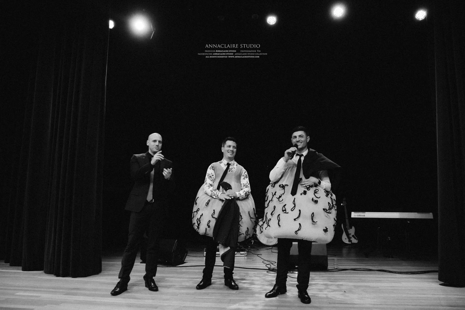 Three men on stage, one holding a microphone, one with a microphone and a jacket draped on his shoulder, and one with a dress with musical notes, under stage lights with a keyboard in the background.