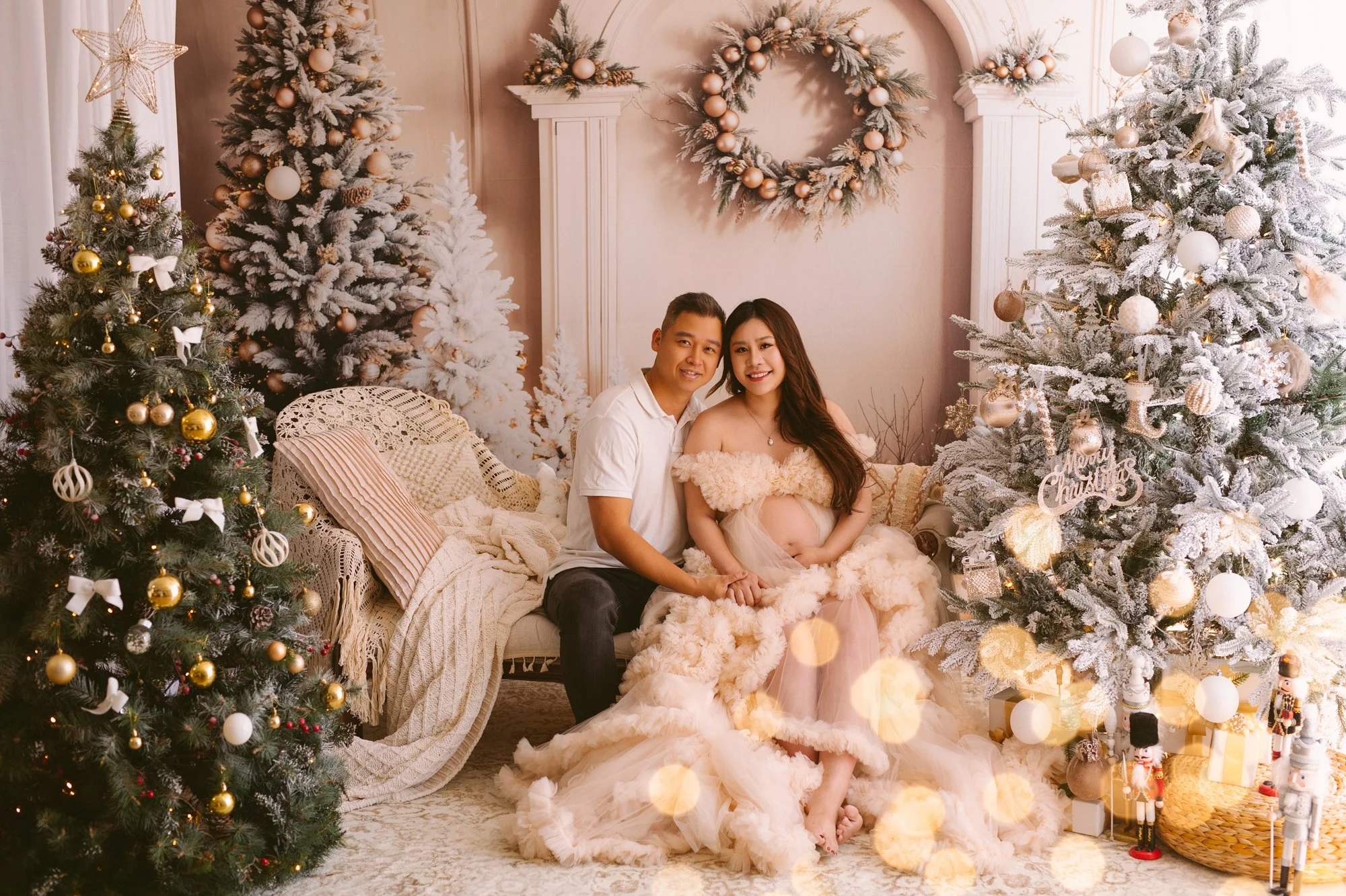 A couple seated on a Christmas-themed sofa, with the woman pregnant, surrounded by Christmas trees and festive decorations, including Christmas ornaments, wreaths, and nutcracker figurines.