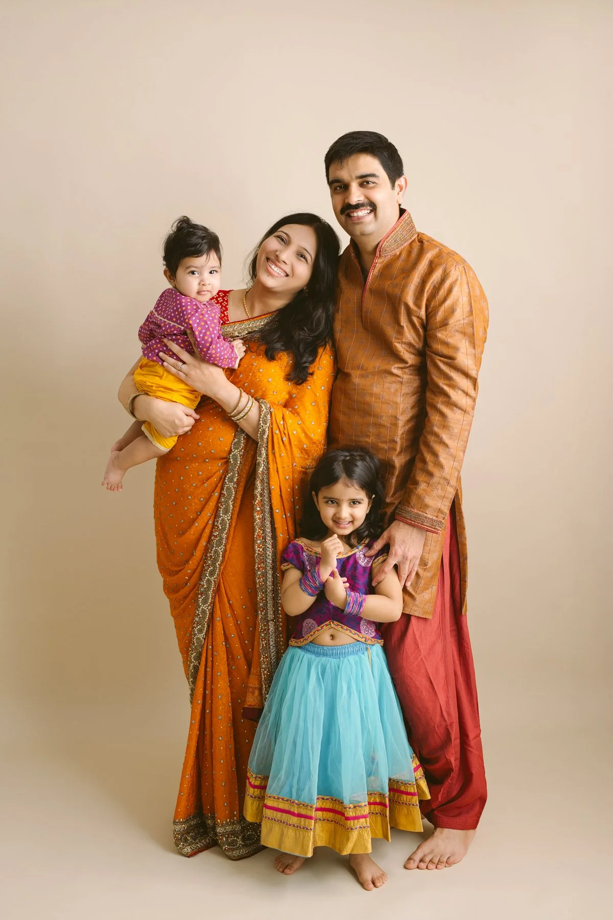 A family of four dressed in traditional Indian clothing posing together against a plain background. The mother holds a young girl, and another girl stands in front of the father, all smiling.